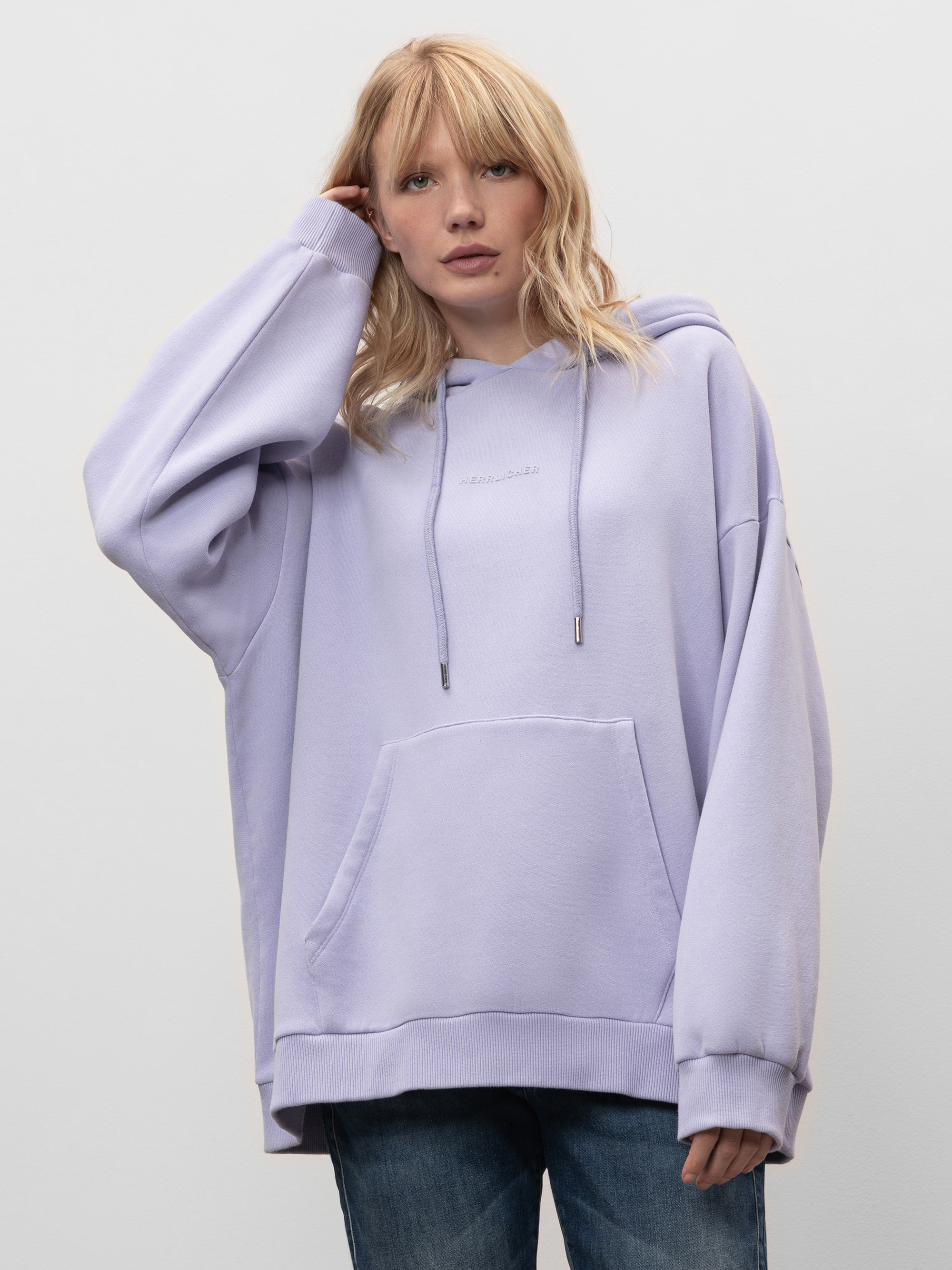 Eine Frau mit blondem Haar, die einen lockeren, hellvioletten Kapuzenpullover und blaue Jeans trägt, steht vor einem schlichten hellgrauen Hintergrund, berührt mit einer Hand ihr Haar und blickt in die Kamera.