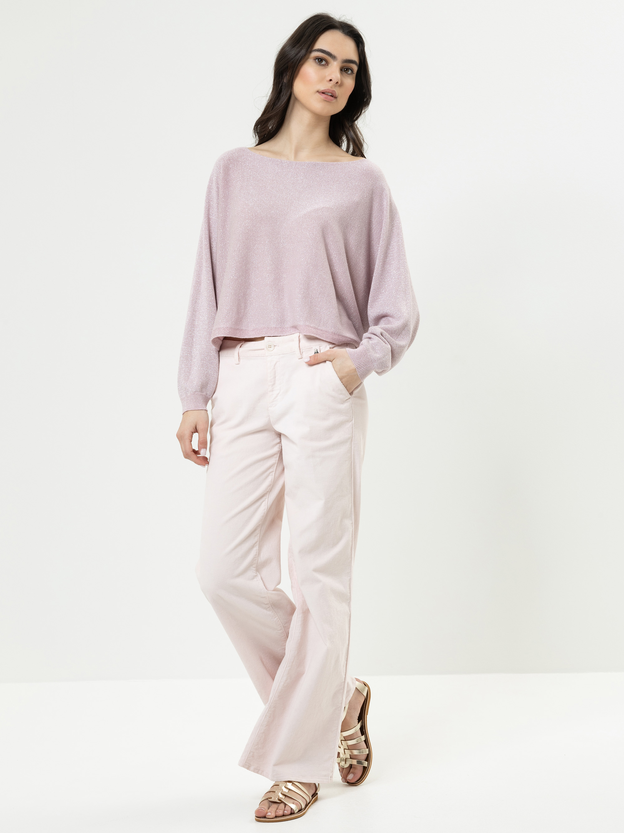 Eine Frau steht vor einem schlichten weißen Hintergrund, trägt ein lockeres, hellviolettes, langärmeliges Oberteil, eine hellrosa, weit geschnittene Hose und goldene Sandalen, eine Hand in der Tasche und einen entspannten Gesichtsausdruck.