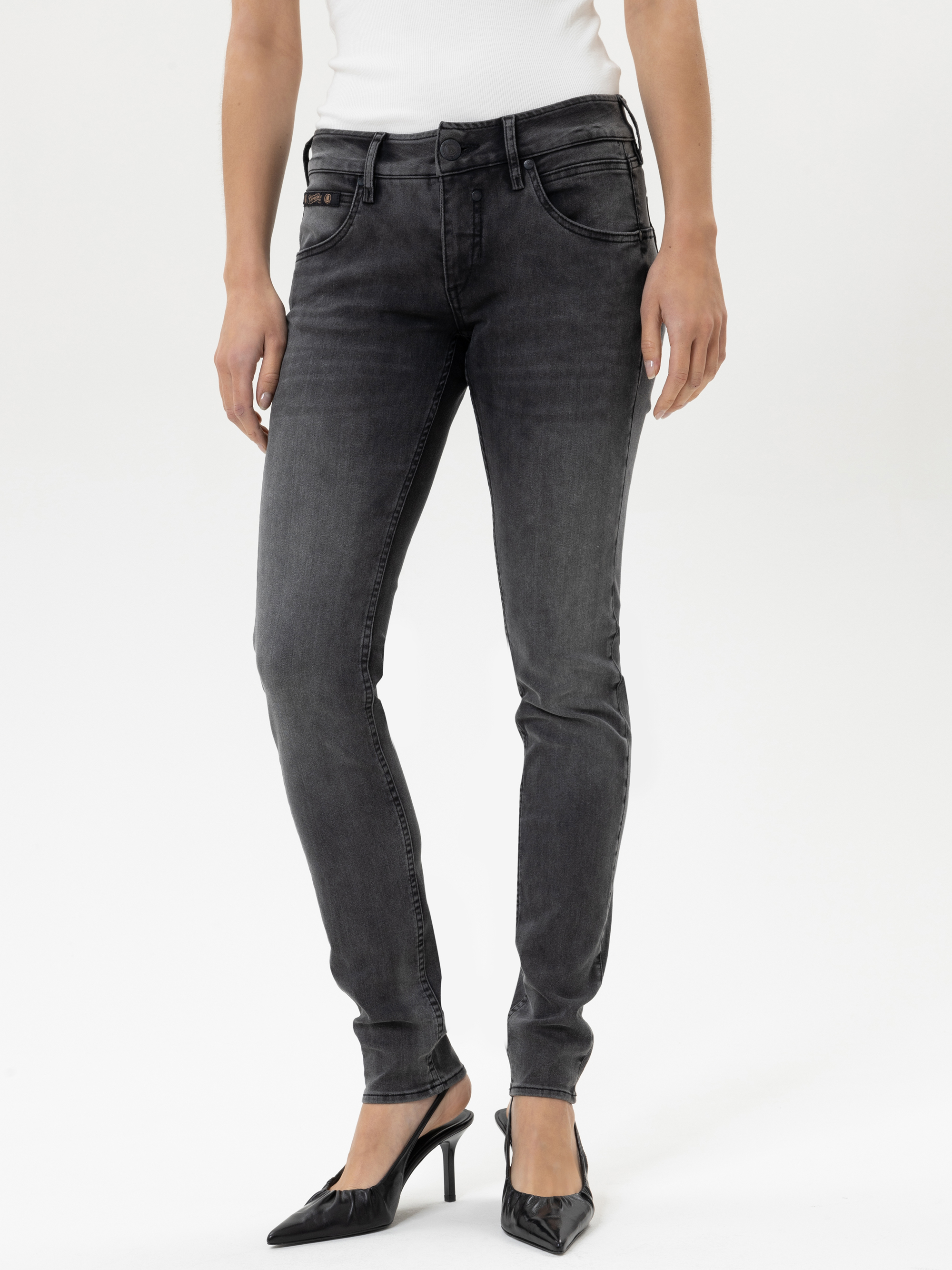 Eine Person, die ein weißes ärmelloses Oberteil, dunkelgraue Röhrenjeans und schwarze spitze Absätze trägt, steht vor einem schlichten weißen Hintergrund.