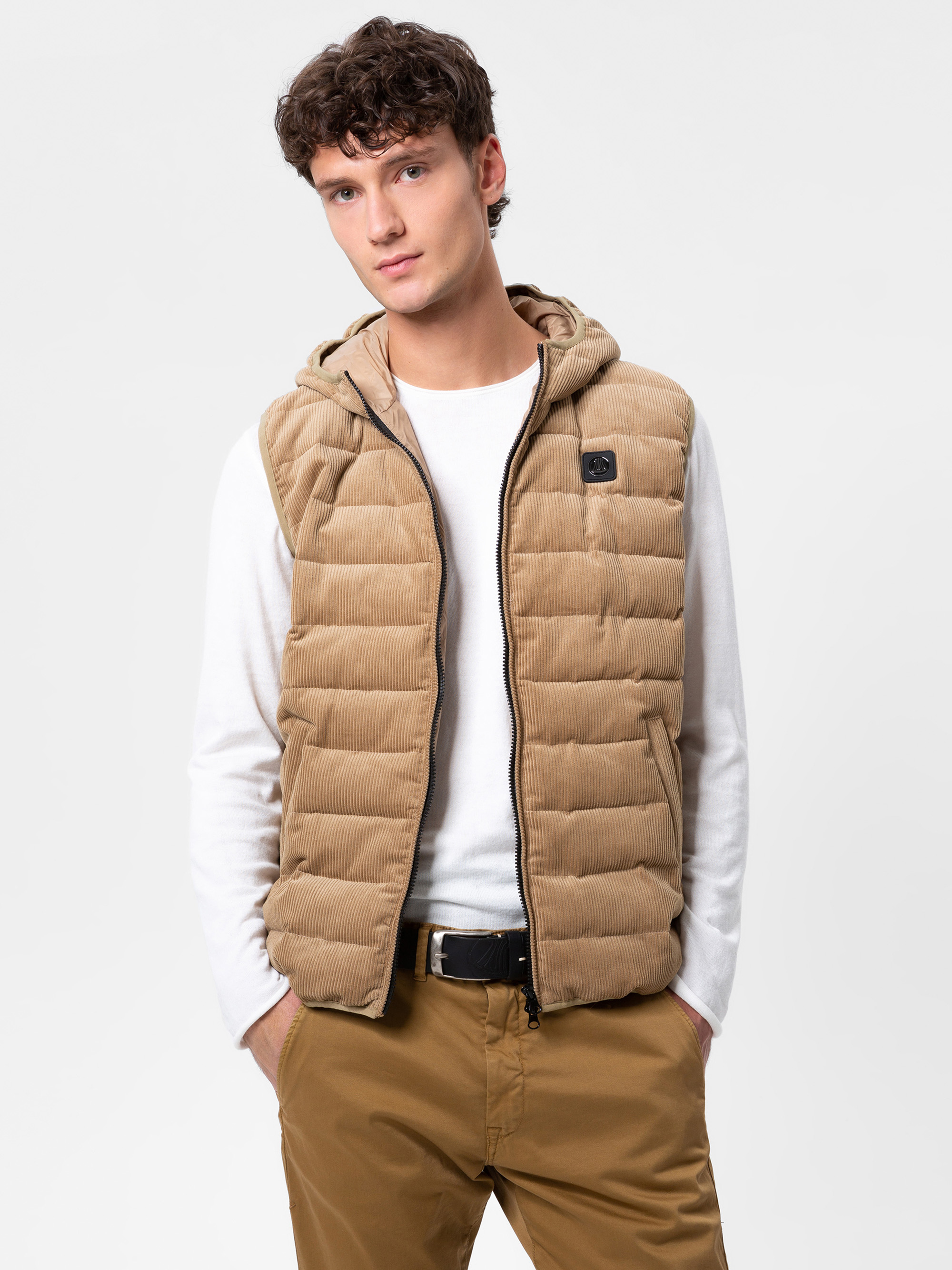 Ein junger Mann, der eine hellbraune Steppweste mit Kapuze über einem weißen langärmeligen Hemd und einer hellbraunen Hose trägt, steht mit den Händen in den Westentaschen und blickt in die Kamera vor einem schlichten weißen Hintergrund.