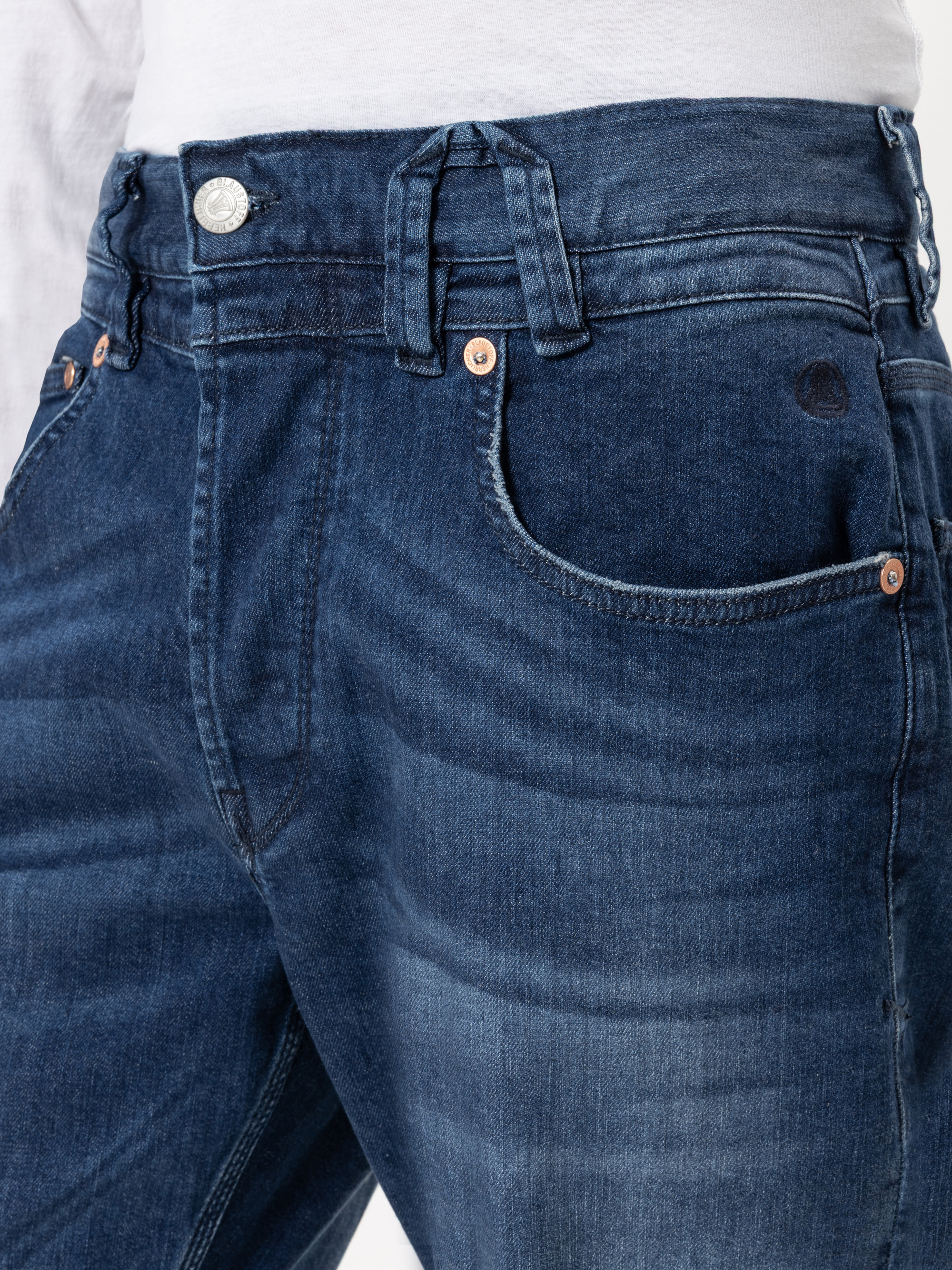 Nahaufnahme einer Person, die eine mittelschwere Bluejeans und ein weißes Hemd trägt. Die Jeans hat Kupfernieten, Gürtelschlaufen und sichtbare Nähte, eine leicht verblasste Textur und einen Knopfverschluss am Bund.