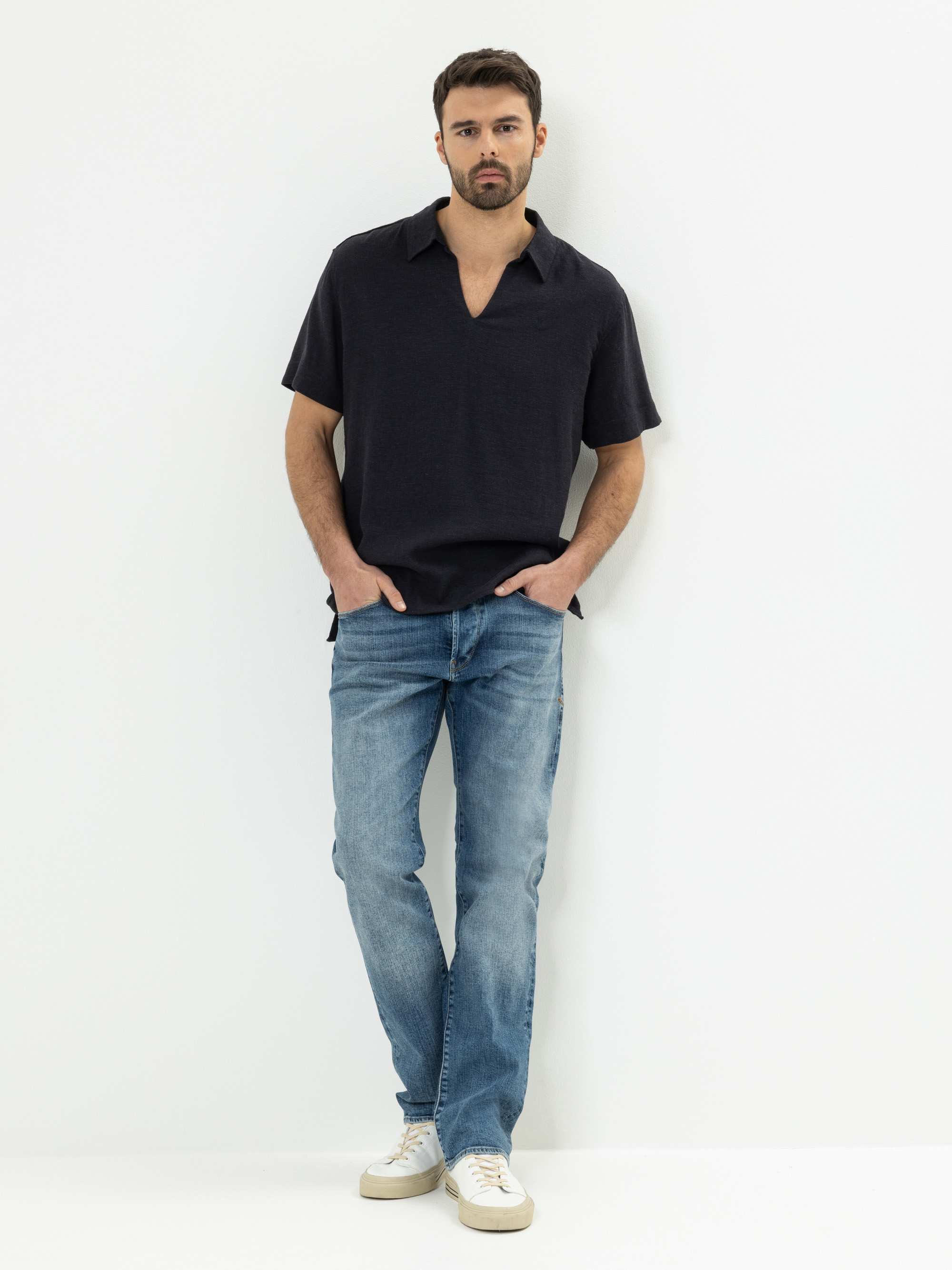 Ein Mann steht vor einem weißen Hintergrund und trägt ein schwarzes kurzärmeliges Hemd, blaue Jeans und weiße Turnschuhe. Er hat die Hände in den Taschen und blickt direkt in die Kamera.