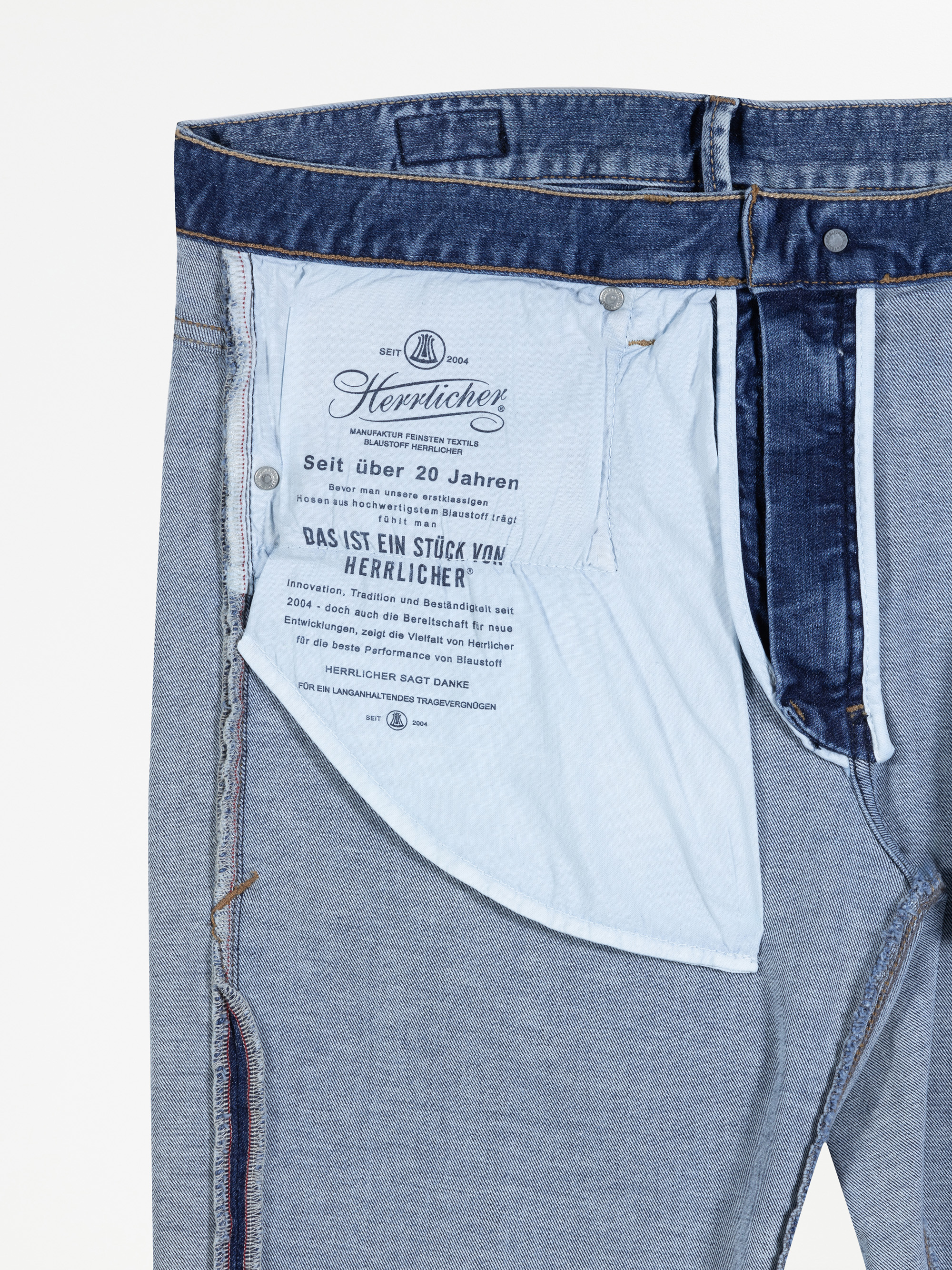 Eine Nahaufnahme einer umgedrehten Bluejeans, die das mit deutschem Text bedruckte Taschenfutter zeigt, einschließlich des Markennamens Herrlicher und Informationen über das Unternehmen. Der Bund der Jeans und die Nähte sind ebenfalls sichtbar.