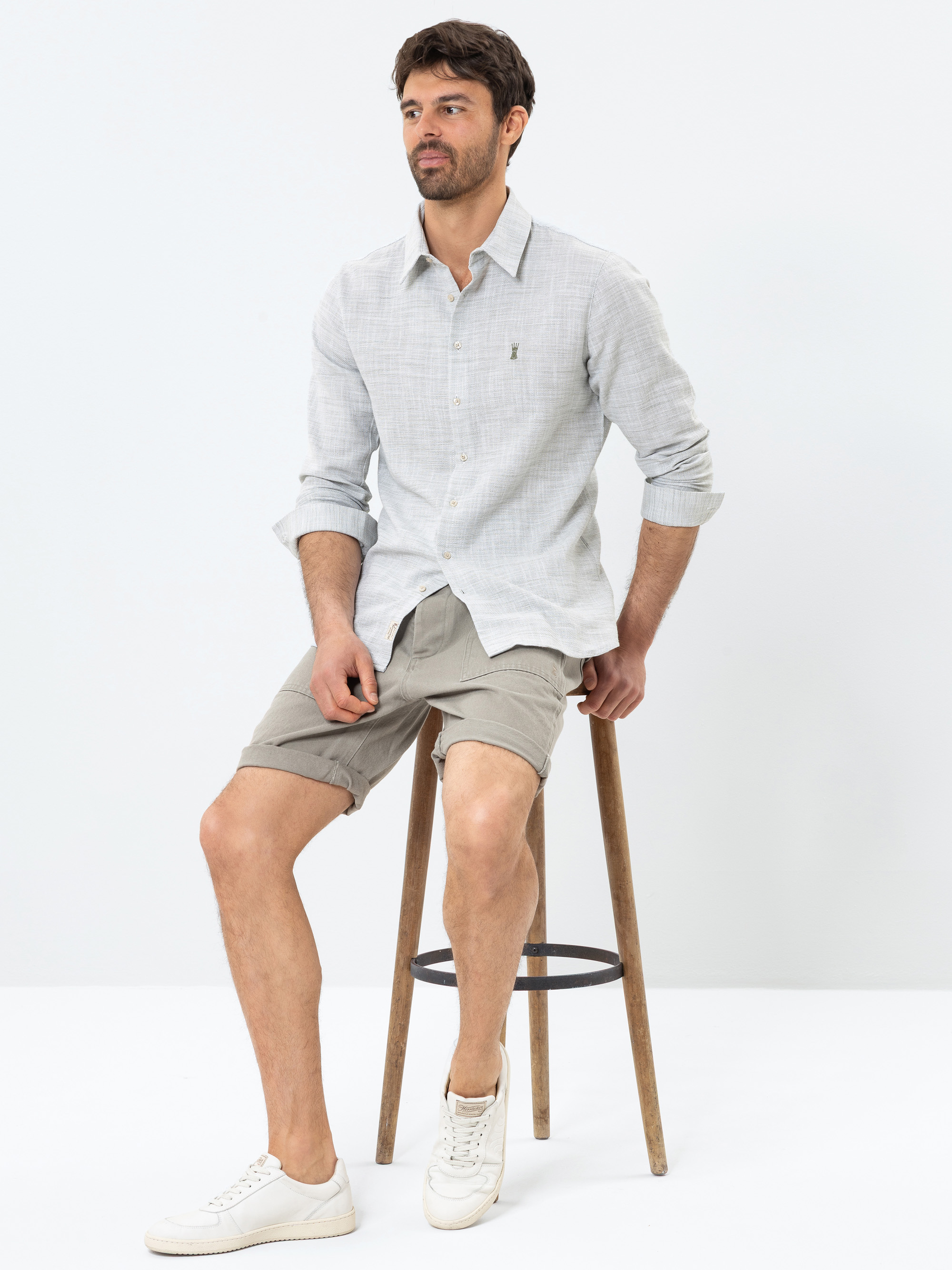 Ein Mann mit kurzen dunklen Haaren und Bart sitzt auf einem Holzschemel und trägt ein hellgraues Button-up-Hemd, beigefarbene Shorts und weiße Turnschuhe vor einem schlichten weißen Hintergrund.