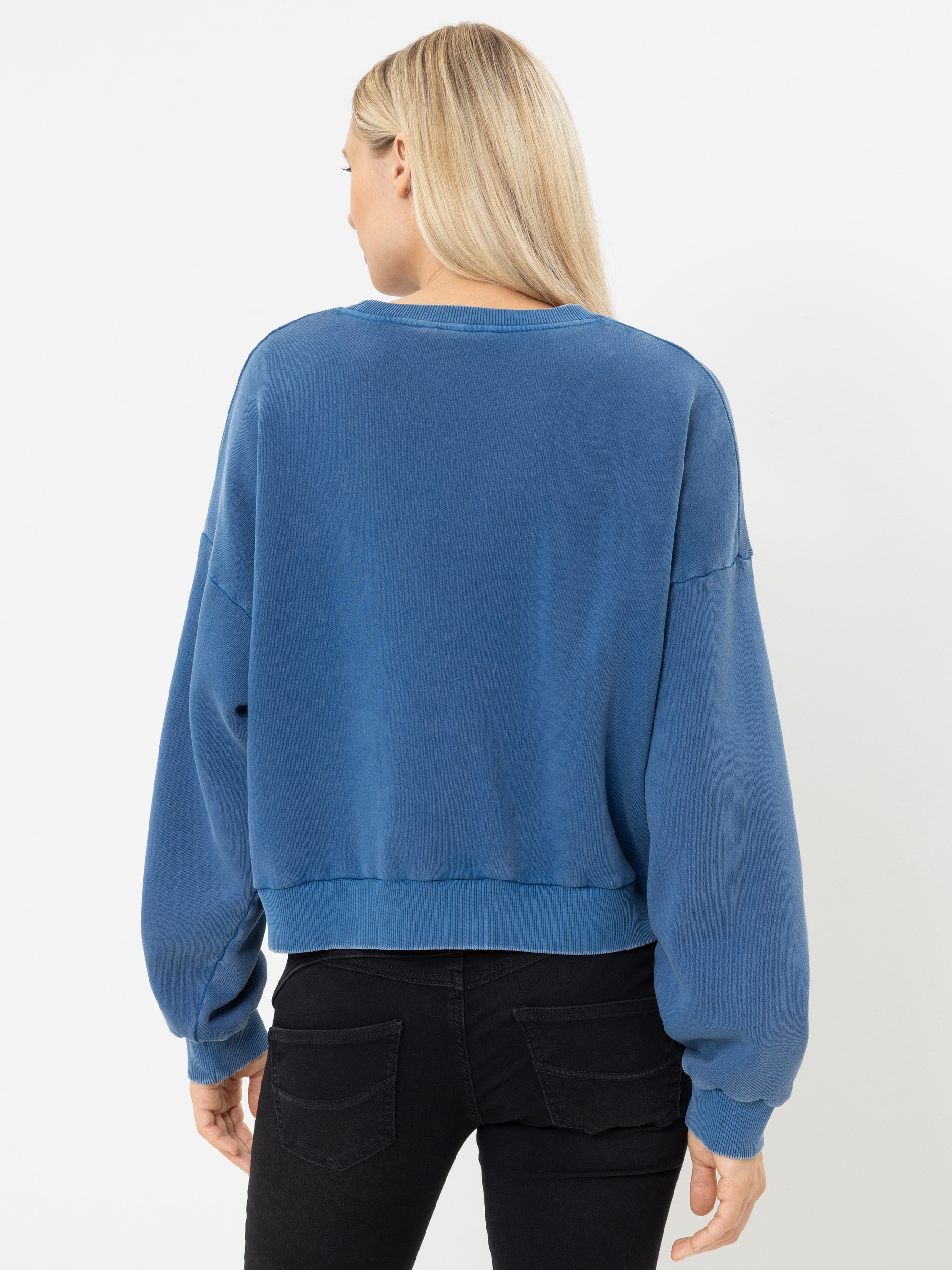 Eine Person mit langen blonden Haaren trägt ein lockeres blaues Sweatshirt und eine schwarze Jeans und steht mit dem Rücken zur Kamera vor einem schlichten weißen Hintergrund.