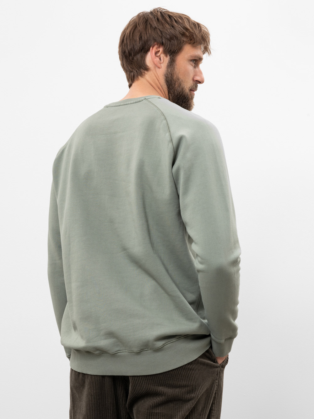 Ein Mann mit hellbraunem Haar und Bart steht seitlich, trägt ein salbeigrünes Sweatshirt und eine braune Cordhose. Er hat die Hände in den Taschen und blickt nach rechts vor einem schlichten weißen Hintergrund.