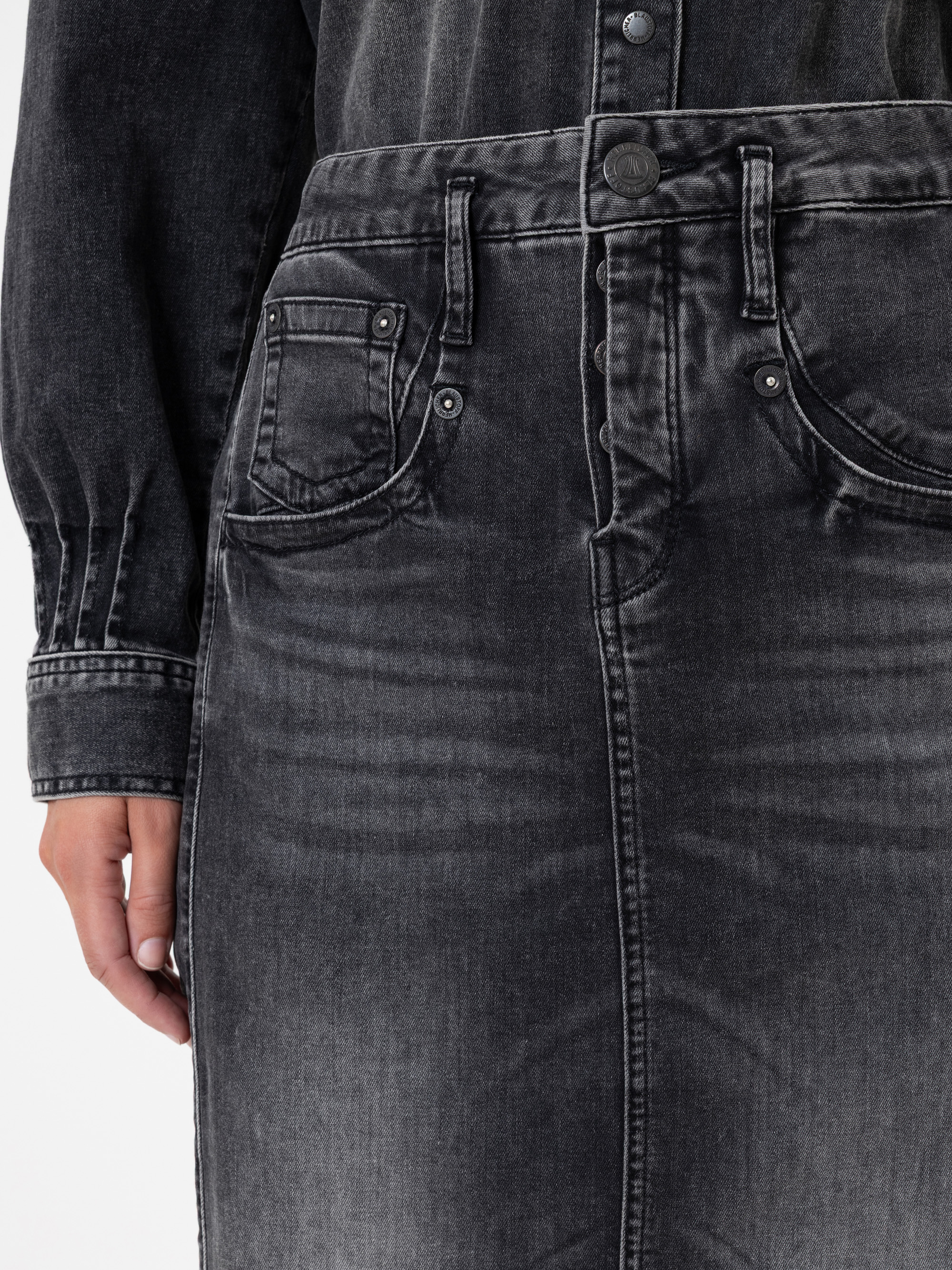 Nahaufnahme einer Person, die einen hochtaillierten, schwarz gewaschenen Jeansrock mit Vordertaschen, Gürtelschlaufen und einem Knopfverschluss trägt, gepaart mit einem passenden Jeanshemd. Nur der untere Teil des Oberkörpers und ein Teil der linken Ha