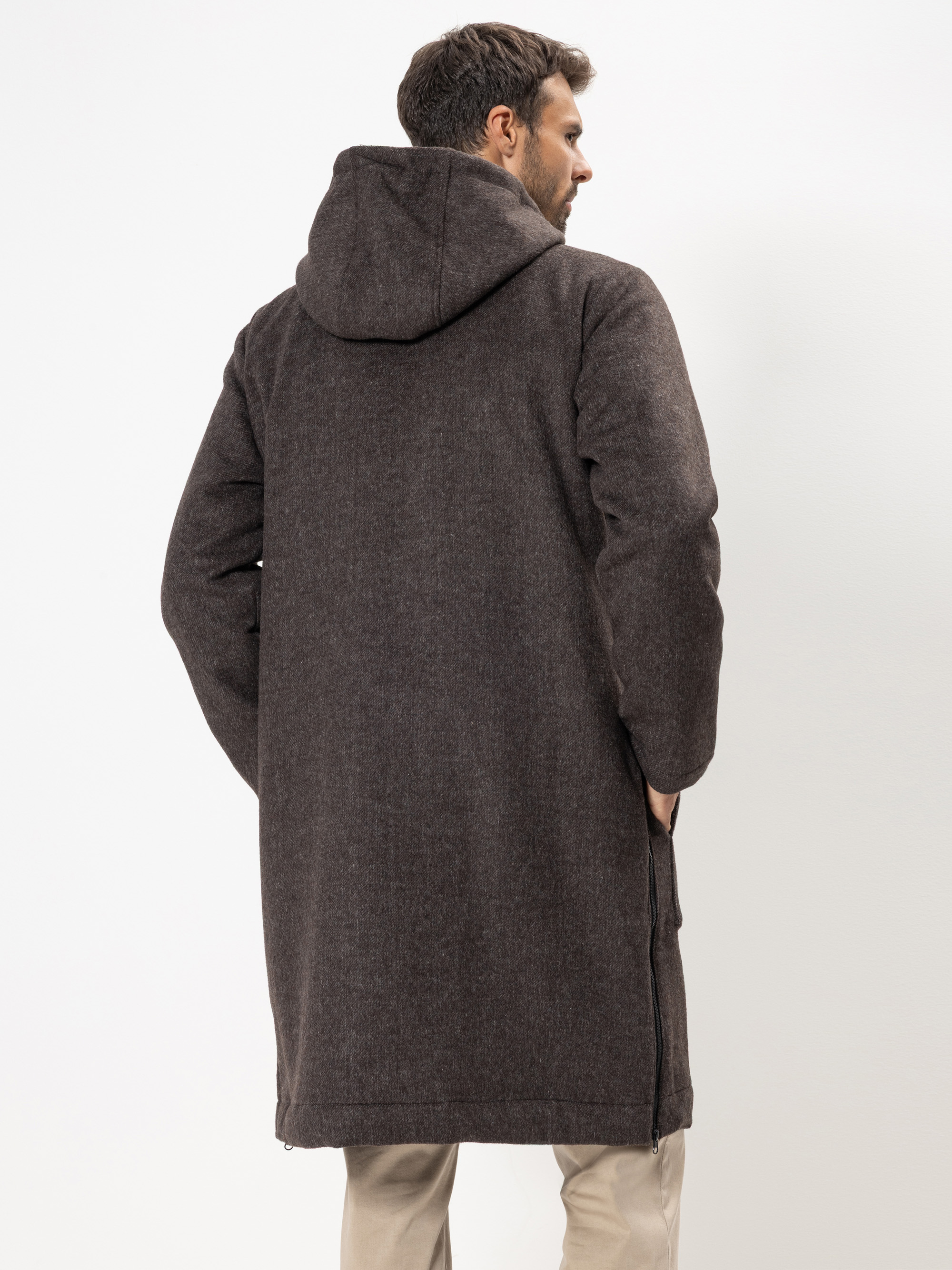 Ein von der Kamera abgewandter Mann trägt einen langen, dunkelbraunen Kapuzenmantel und eine helle Hose und steht vor einem schlichten weißen Hintergrund.
