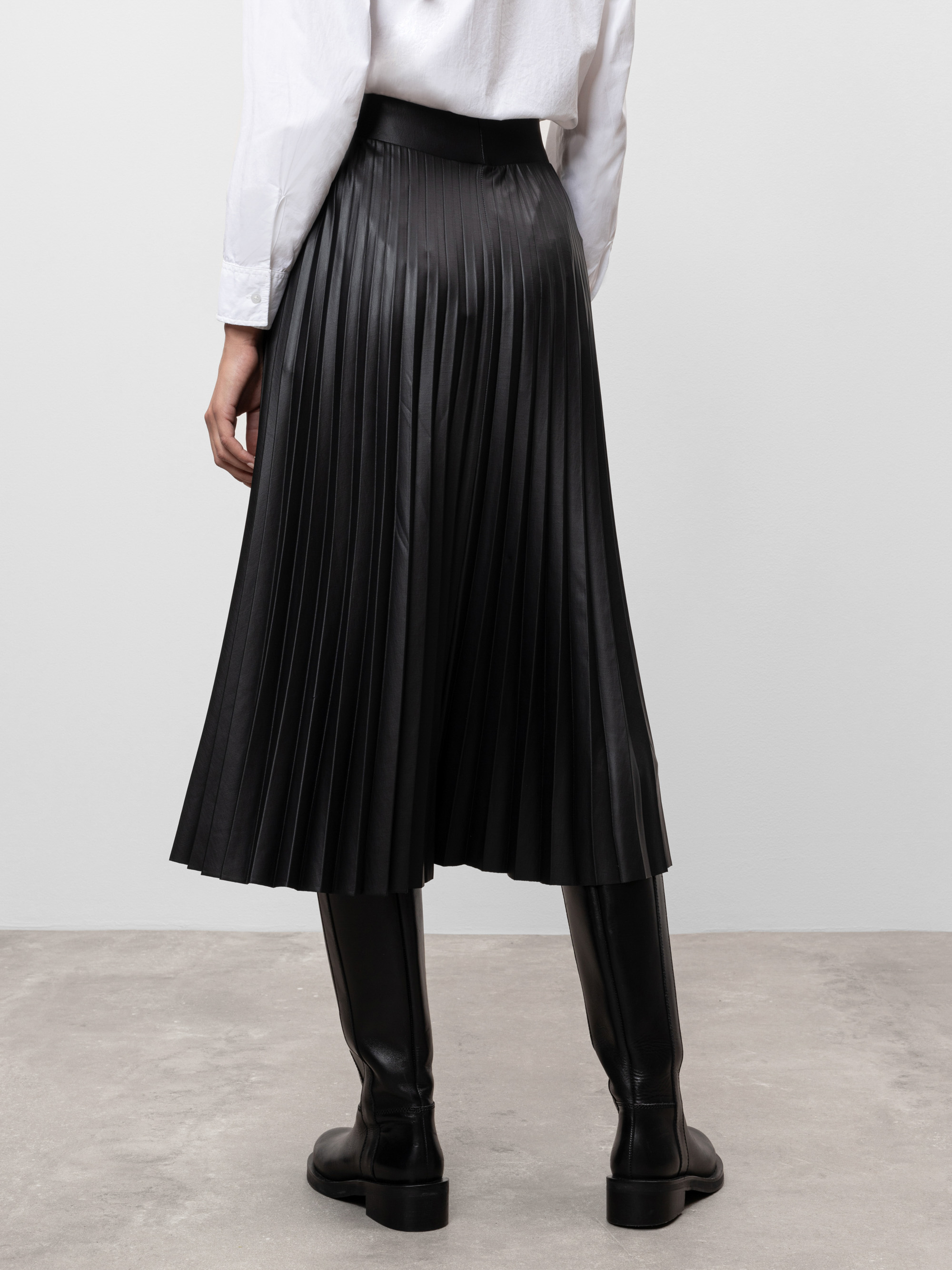 Eine Person steht auf einem grauen Boden und trägt ein weißes langärmeliges Hemd, einen schwarzen Midi-Faltenrock und schwarze kniehohe Stiefel. Das Bild zeigt das Outfit von hinten.