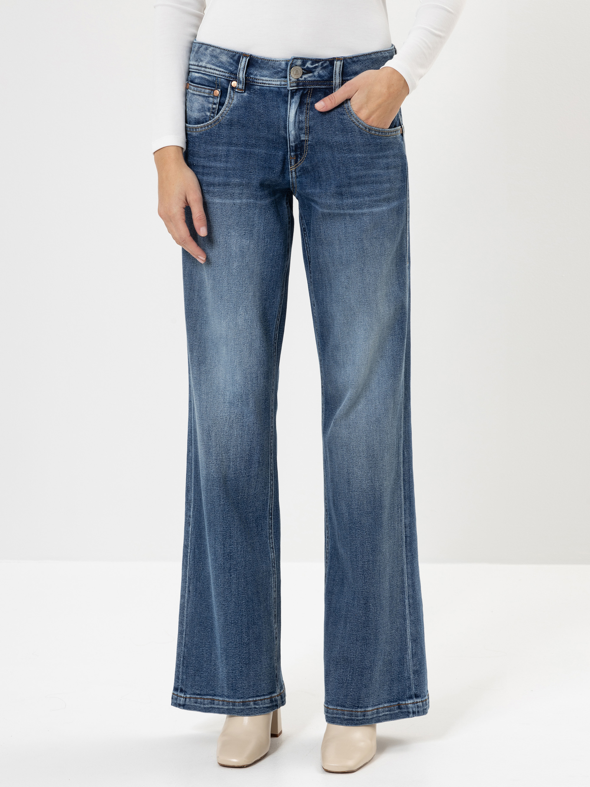 Eine Person, die eine blaue Jeans mit hoher Taille und weitem Bein, ein weißes langärmeliges Oberteil und beigefarbene Schuhe mit Absätzen trägt, steht vor einem schlichten weißen Hintergrund.