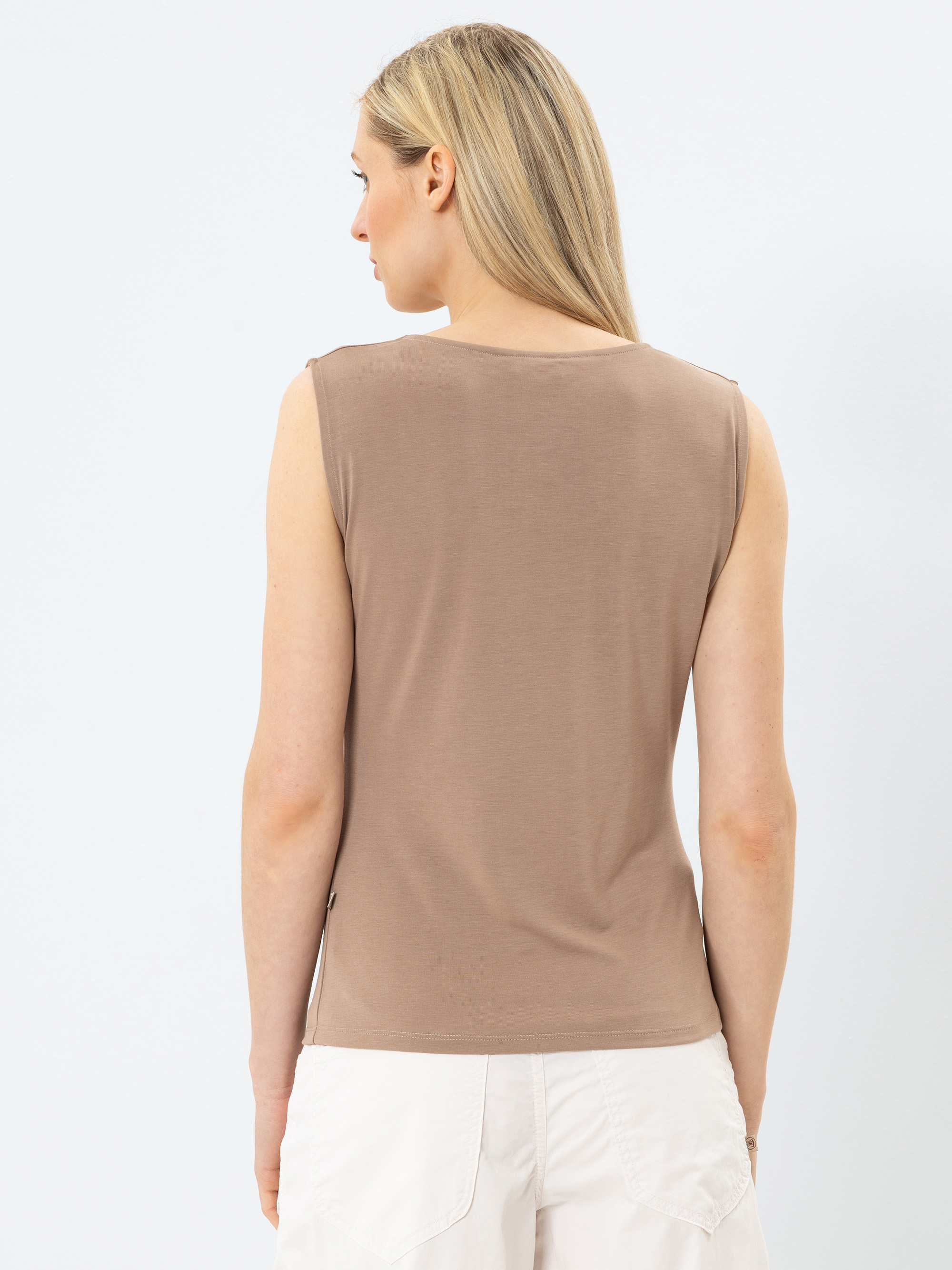 Eine Frau mit langen blonden Haaren ist von hinten zu sehen. Sie trägt ein ärmelloses taupefarbenes Oberteil und weiße Shorts und steht vor einem schlichten hellen Hintergrund.