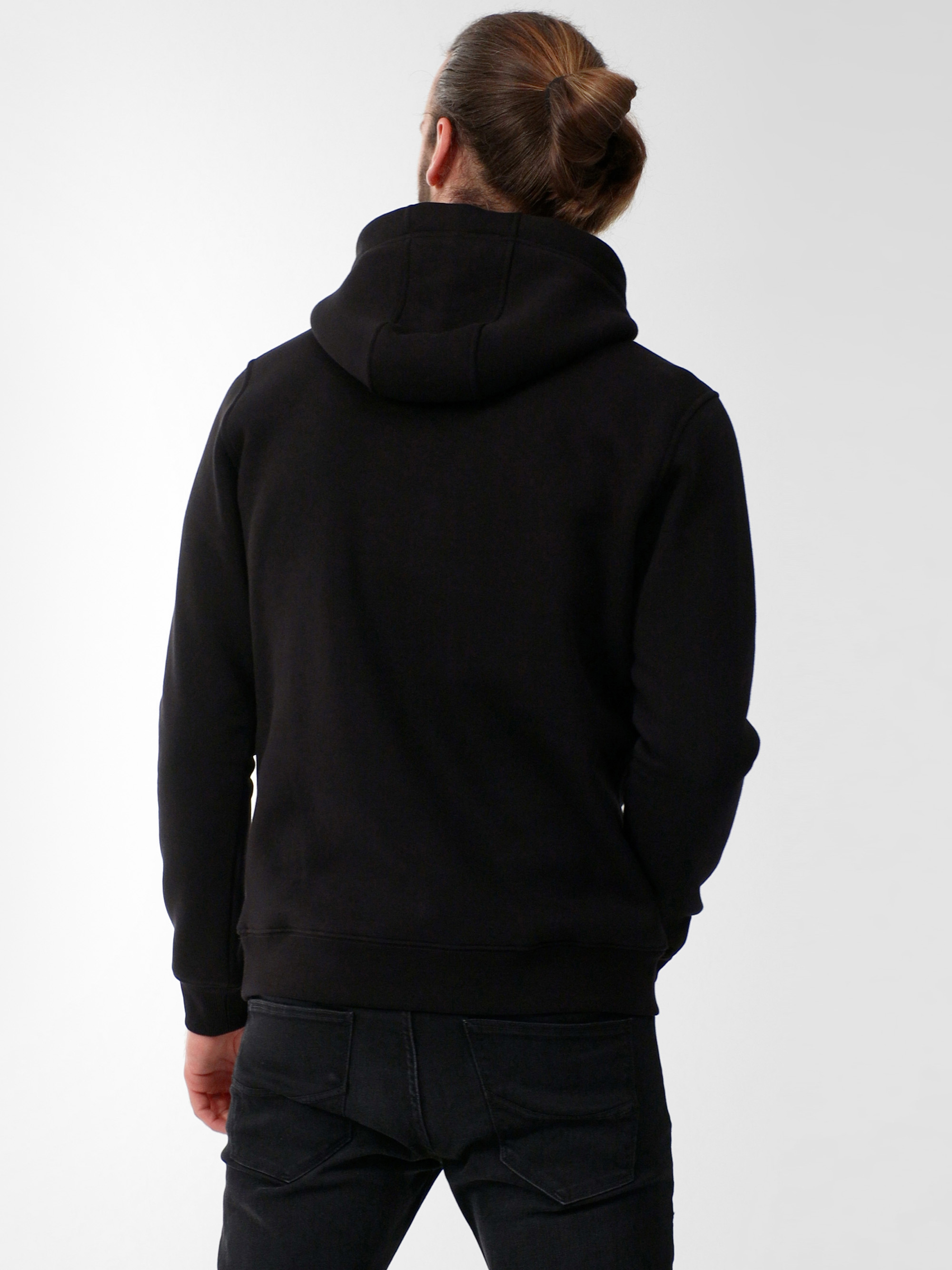 Eine Person mit langen Haaren, die zu einem Dutt gebunden sind, trägt einen schwarzen Kapuzenpullover und schwarze Jeans und steht mit dem Rücken zur Kamera vor einem schlichten hellgrauen Hintergrund.