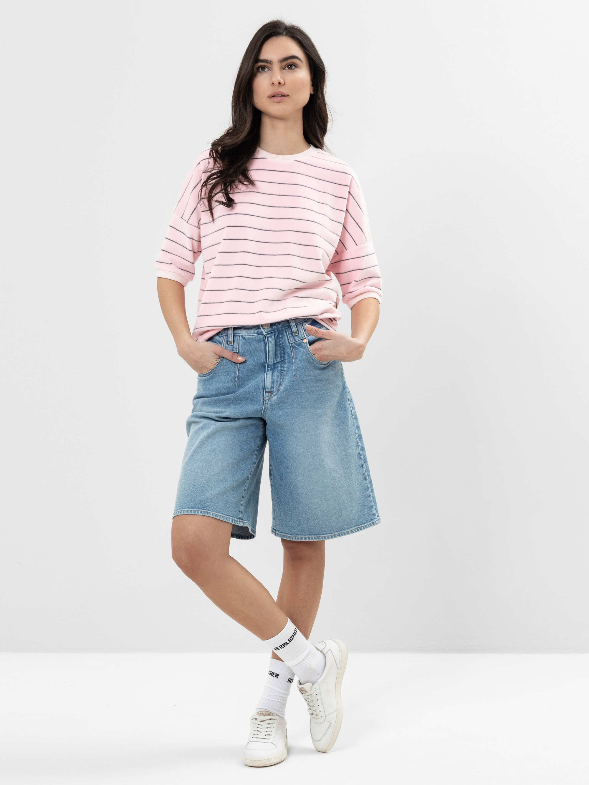 Eine Frau steht vor einem schlichten weißen Hintergrund und trägt ein hellrosa, langärmeliges gestreiftes Hemd, blaue knielange Jeans-Shorts, weiße Socken und weiße Turnschuhe. Sie hat langes dunkles Haar und eine entspannte, selbstbewusste Pose.