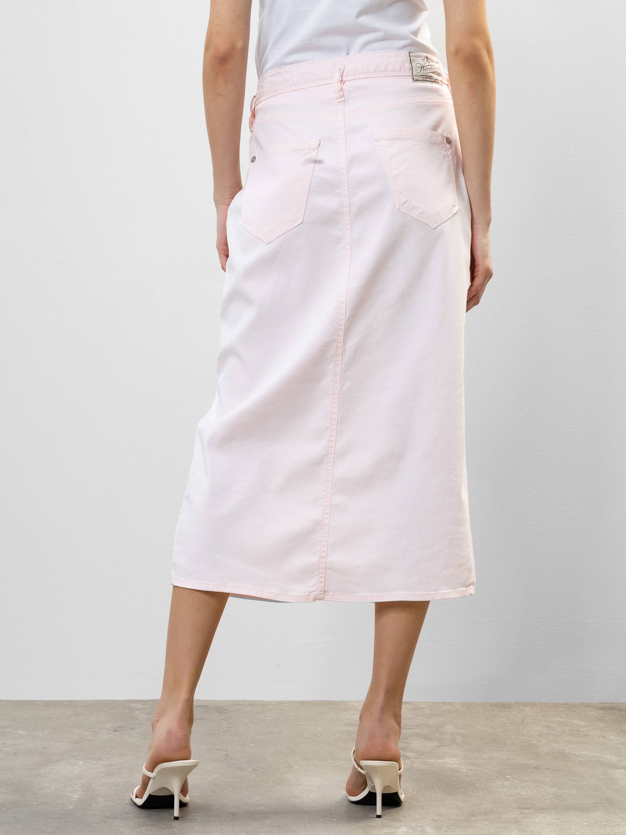 Eine Person, die ein weißes Oberteil, einen hellrosa Jeans-Midirock mit Gesäßtaschen und weiße hochhackige Sandalen trägt, steht auf einem grauen Boden und schaut von der Kamera weg.