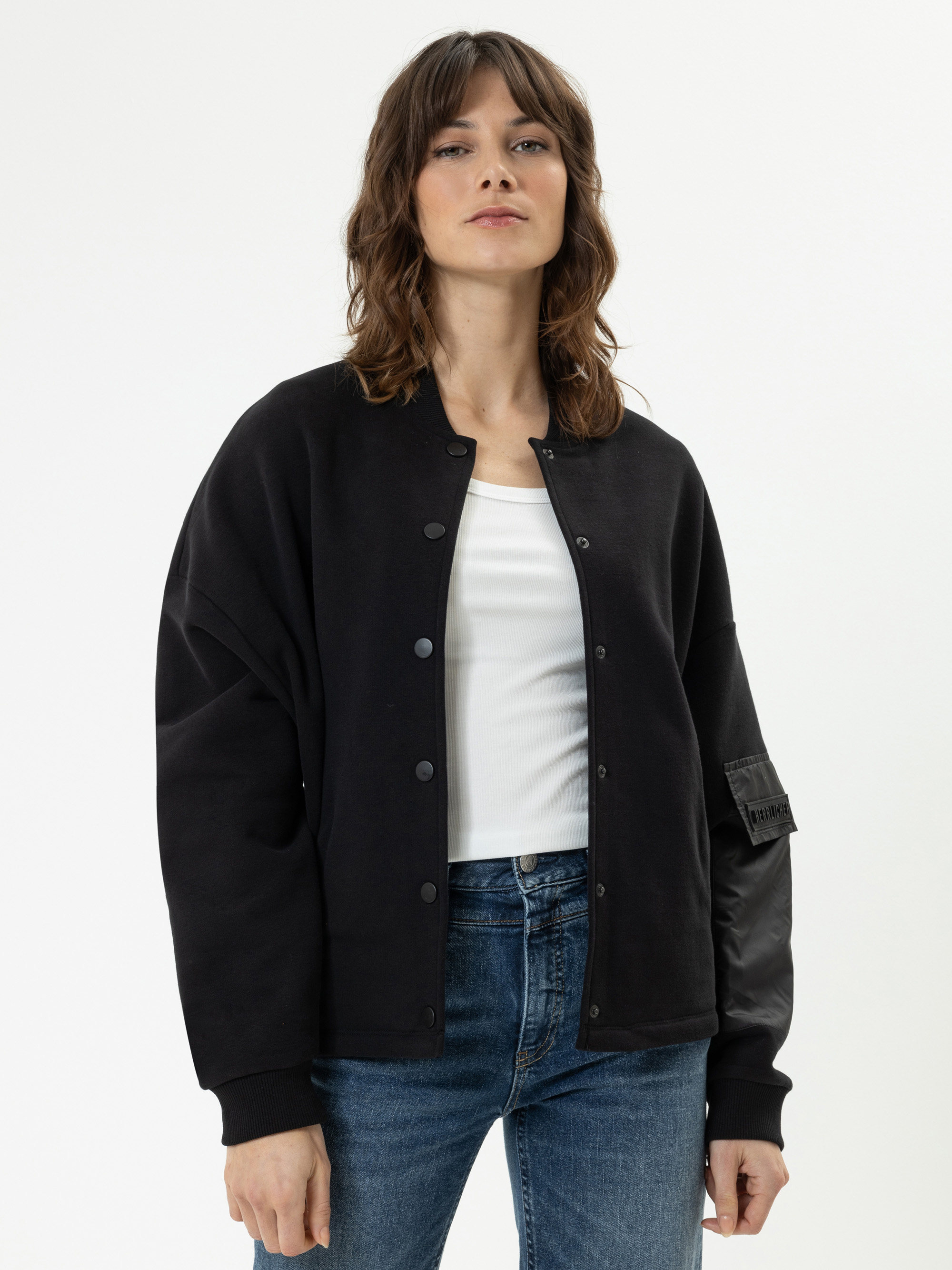 Eine Frau mit gewelltem braunem Haar trägt eine schwarze Button-up-Jacke über einem weißen Oberteil und blaue Jeans. Sie steht vor einem schlichten weißen Hintergrund und blickt selbstbewusst in die Kamera.