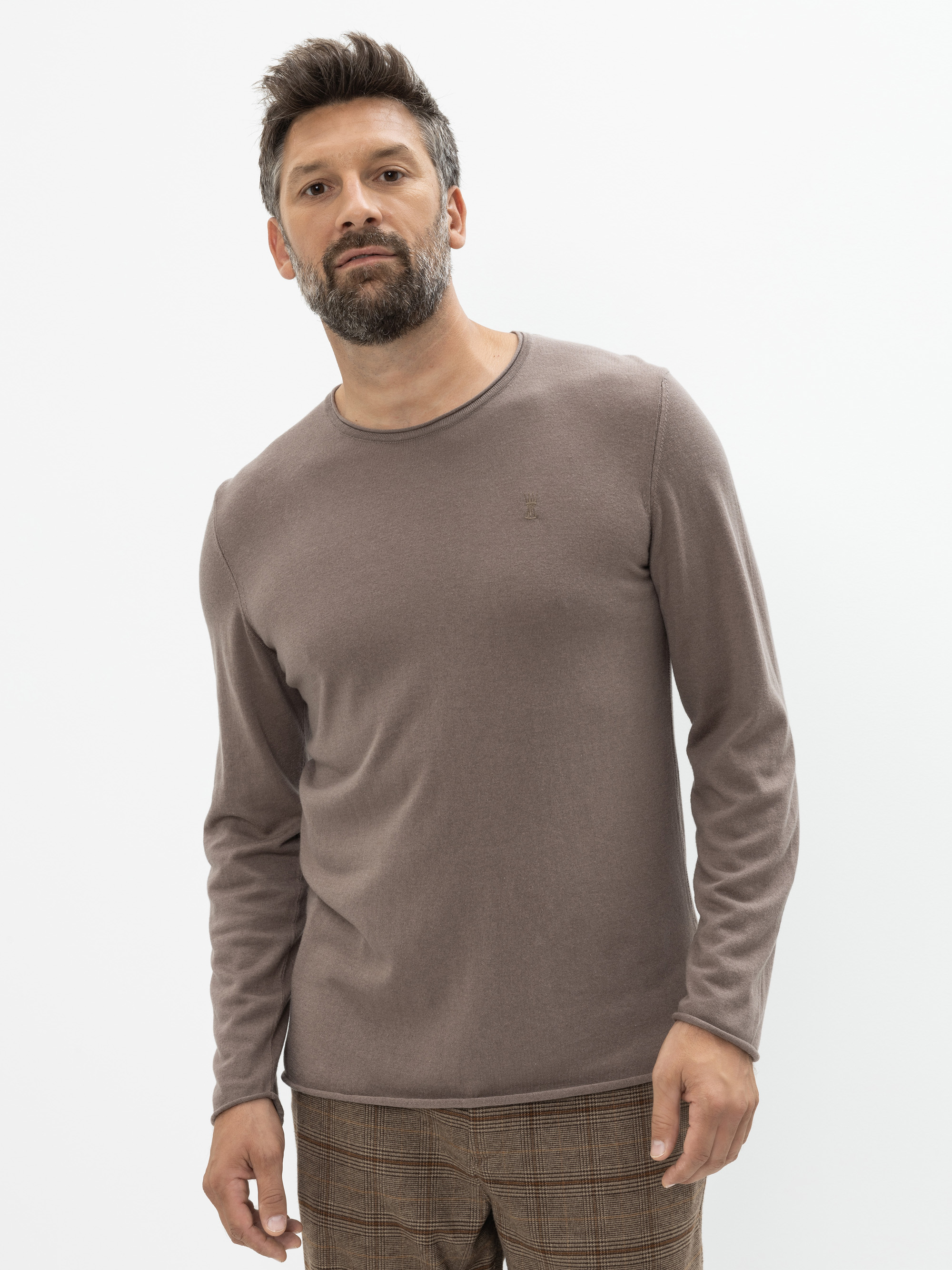 Ein Mann mit kurzen dunklen Haaren und einem Bart trägt einen langärmeligen lilafarbenen Pullover und eine karierte braune Hose und steht vor einem schlichten weißen Hintergrund.