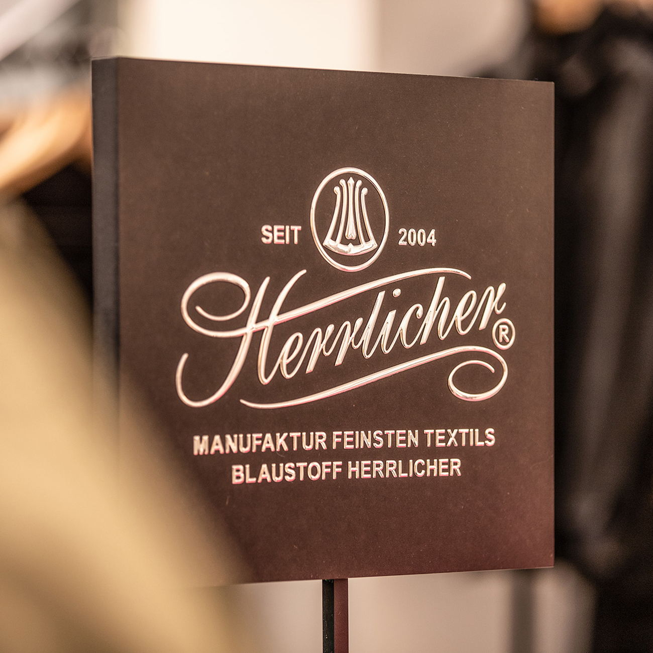 Ein dunkles Schild zeigt das Logo "Herrlicher" mit dem Text "Seit 2004", "Manufaktur Feinsten Textils" und "Blaustoff Herrlicher" in eleganten Schriftarten, was auf einen 2004 gegründeten Textilhersteller hinweist.