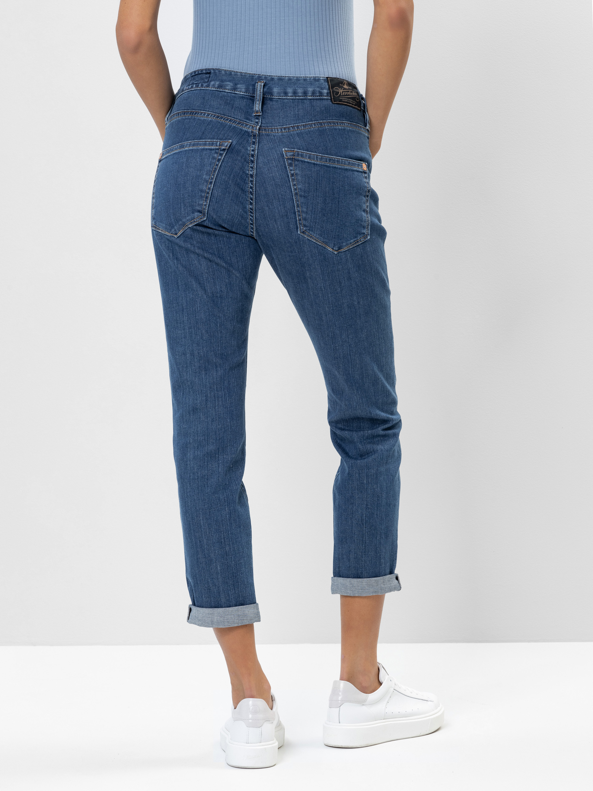 Person in blauen Jeans mit Bündchen und weißen Turnschuhen, die mit dem Rücken zur Kamera vor einem schlichten hellen Hintergrund steht. Der Oberkörper ist teilweise sichtbar, bekleidet mit einem hellblauen gerippten Oberteil.