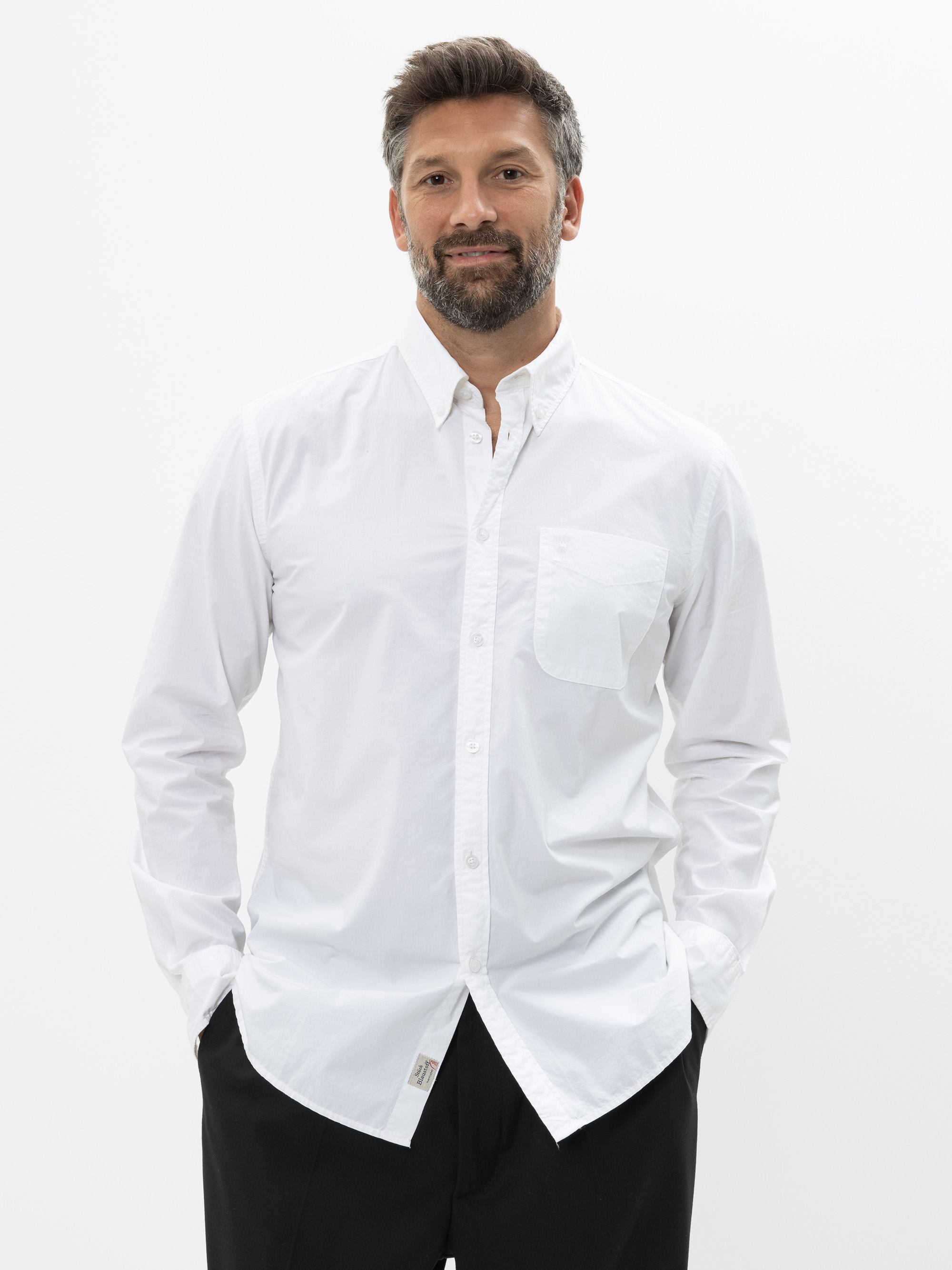 Vor einem schlichten weißen Hintergrund steht ein Mann mit kurzen braunen Haaren und Bart, der ein weißes Button-up-Hemd und eine schwarze Hose trägt, die Hände in den Taschen und ein leichtes Lächeln im Gesicht.