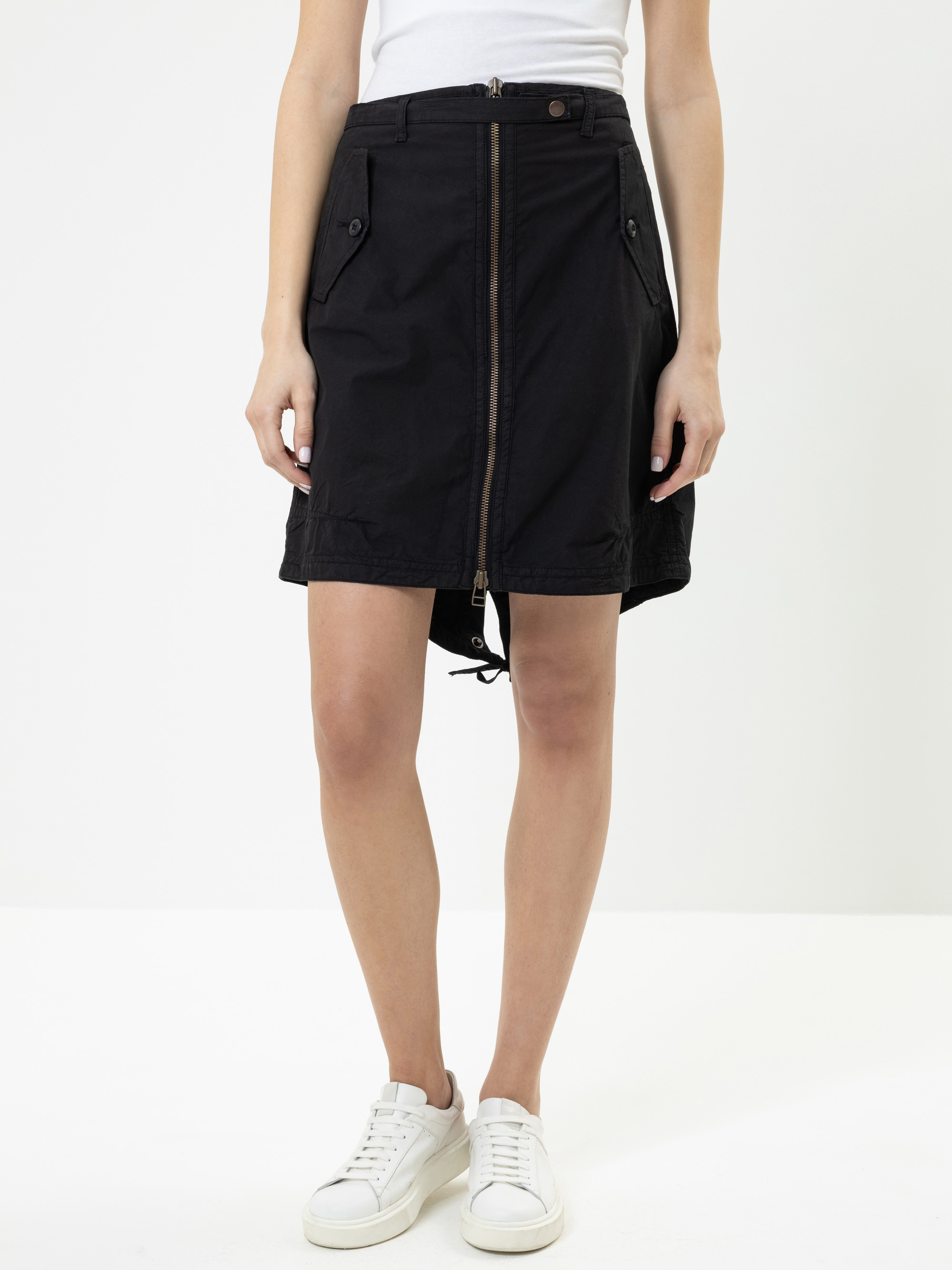 Eine Person, die ein weißes Oberteil, einen schwarzen Rock mit Reißverschluss, Taschen und Knöpfen sowie weiße Turnschuhe trägt, steht vor einem schlichten weißen Hintergrund.