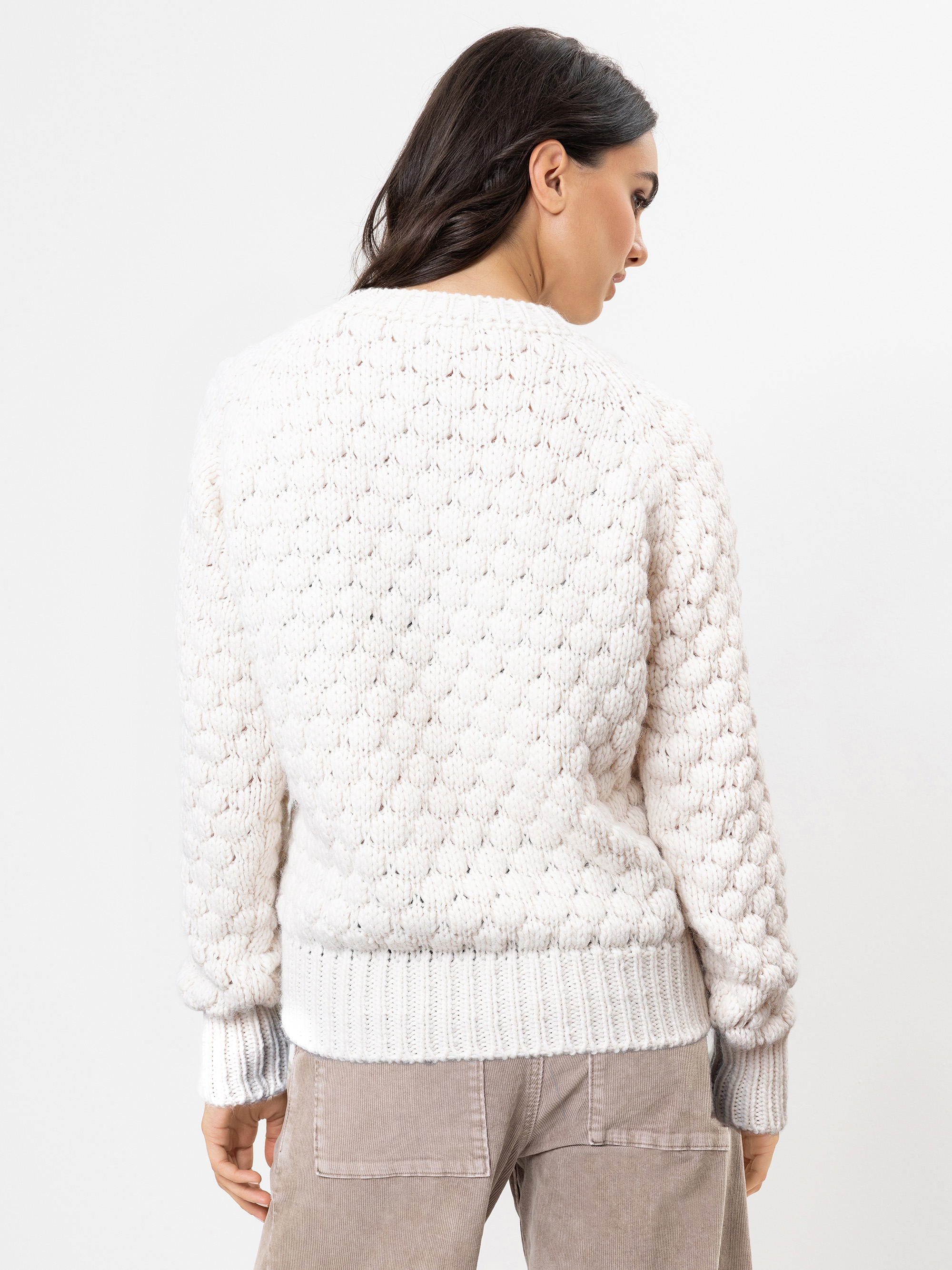 Eine Frau mit dunklem Haar trägt einen strukturierten, cremefarbenen Strickpullover und eine hellbeige Hose. Sie blickt von der Kamera weg und zeigt die Rückseite ihres Outfits vor einem schlichten weißen Hintergrund.