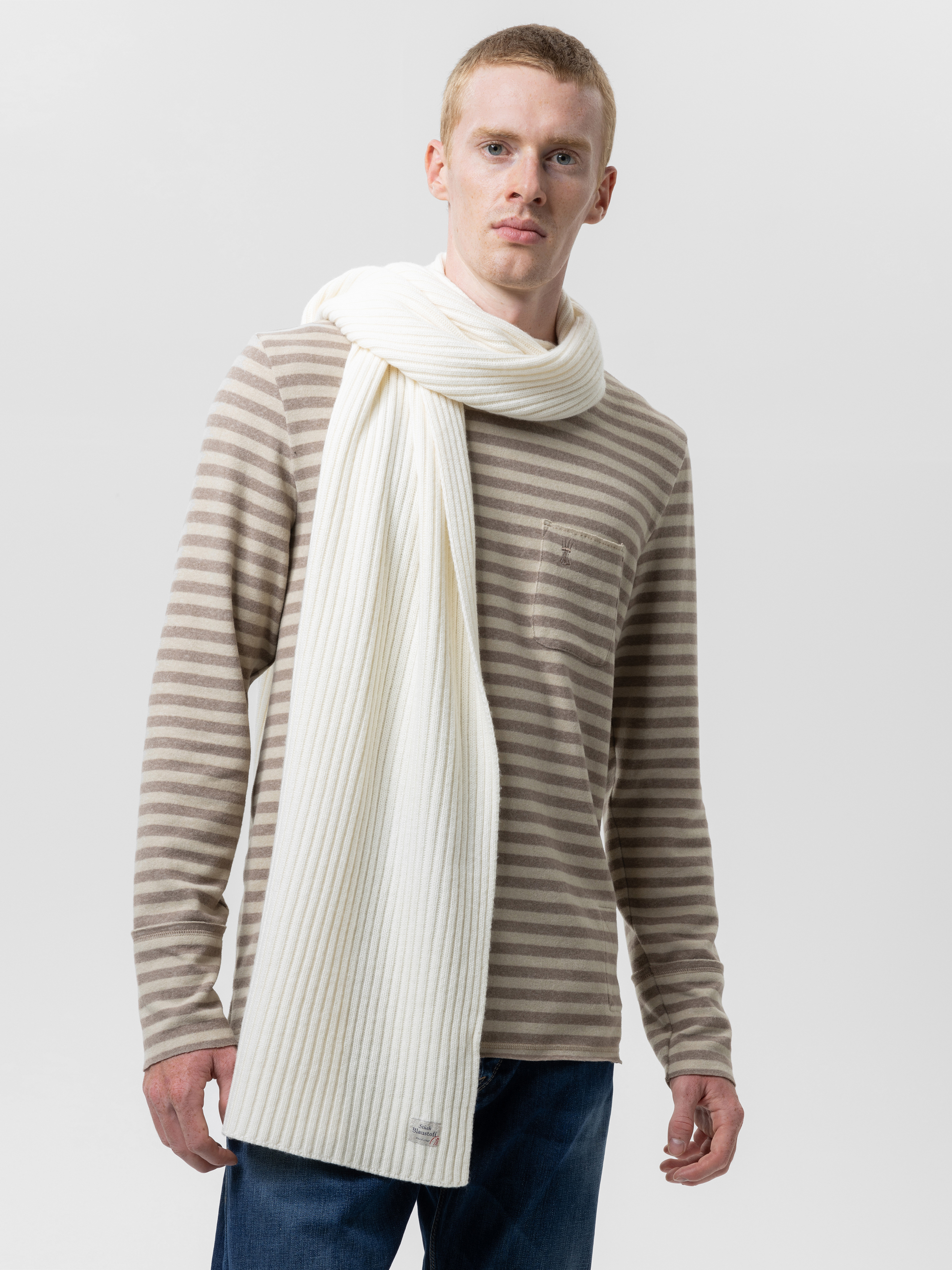 Ein Mann, der ein beige-weiß gestreiftes, langärmeliges Hemd, blaue Jeans und einen langen, gerippten, cremefarbenen Schal trägt, steht vor einem schlichten, hellen Hintergrund.