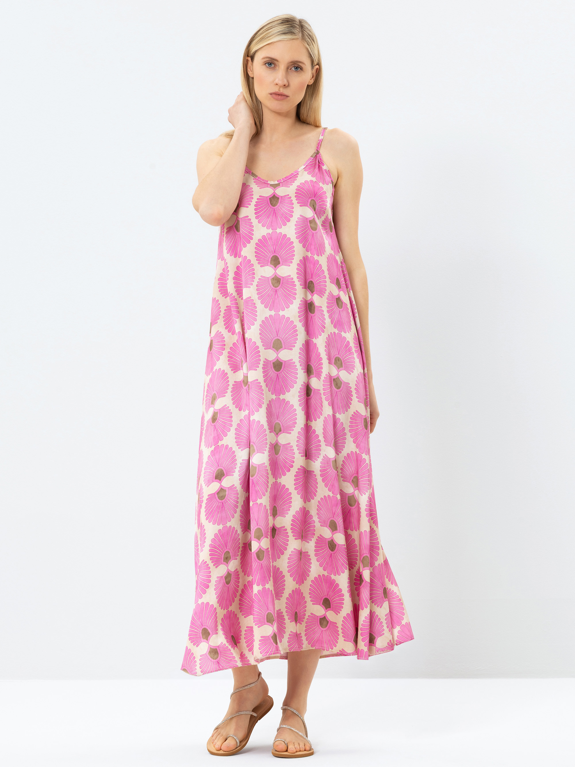 Eine Frau steht vor einem schlichten weißen Hintergrund und trägt ein ärmelloses, rosa Maxikleid mit einem auffälligen Blumenmuster und hellbraune Sandalen. Sie hat glattes blondes Haar und einen neutralen Gesichtsausdruck, wobei eine Hand ihr Gesicht