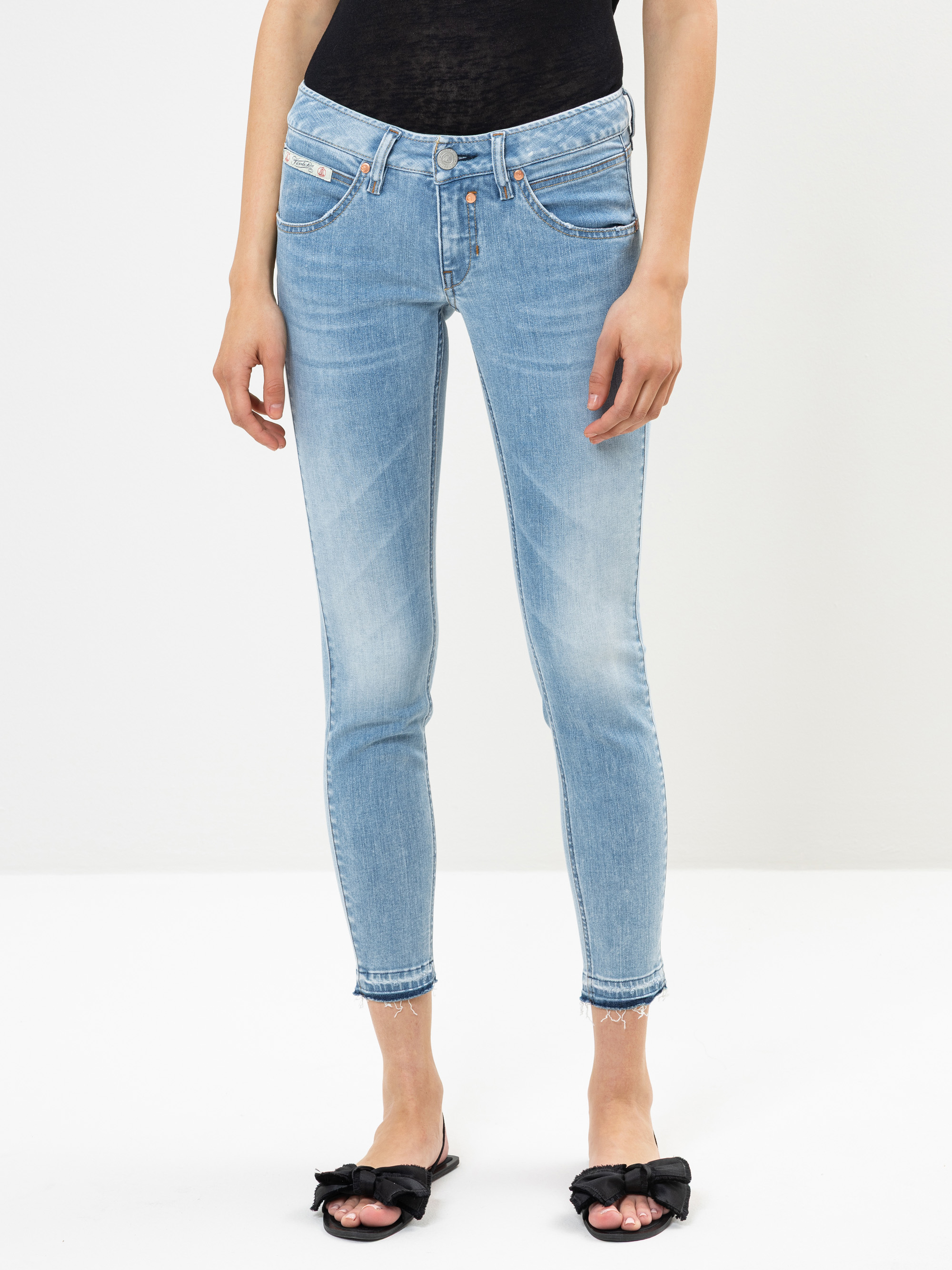 Eine Person, die hellblaue Röhrenjeans mit ausgefransten Säumen, ein schwarzes Oberteil und schwarze offene Sandalen mit Schleifendetails trägt, steht vor einem schlichten weißen Hintergrund.