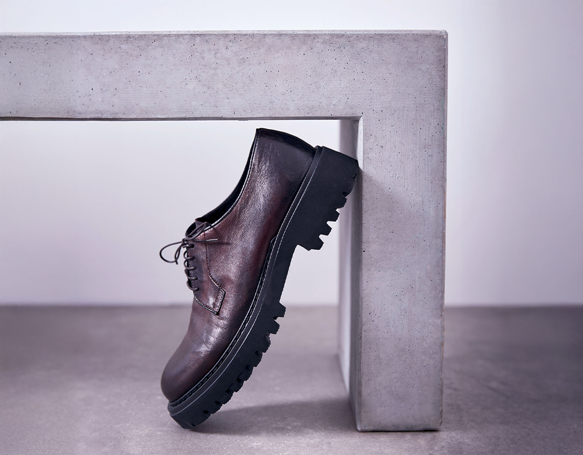 Ein brauner Lederschnürschuh mit klobiger schwarzer Sohle lehnt an der Innenecke einer modernen, hellgrauen Betonbank auf einem glatten Boden.