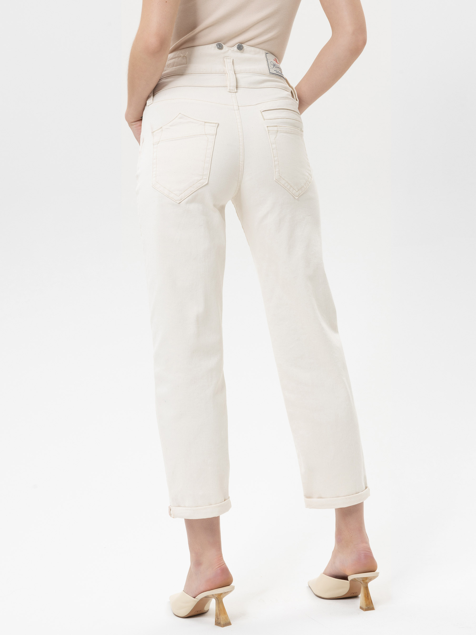 Eine Person steht mit dem Rücken zur Kamera, trägt eine hellbeige, hochgekrempelte Jeans und beigefarbene Pantoletten mit Absatz vor einem schlichten weißen Hintergrund.