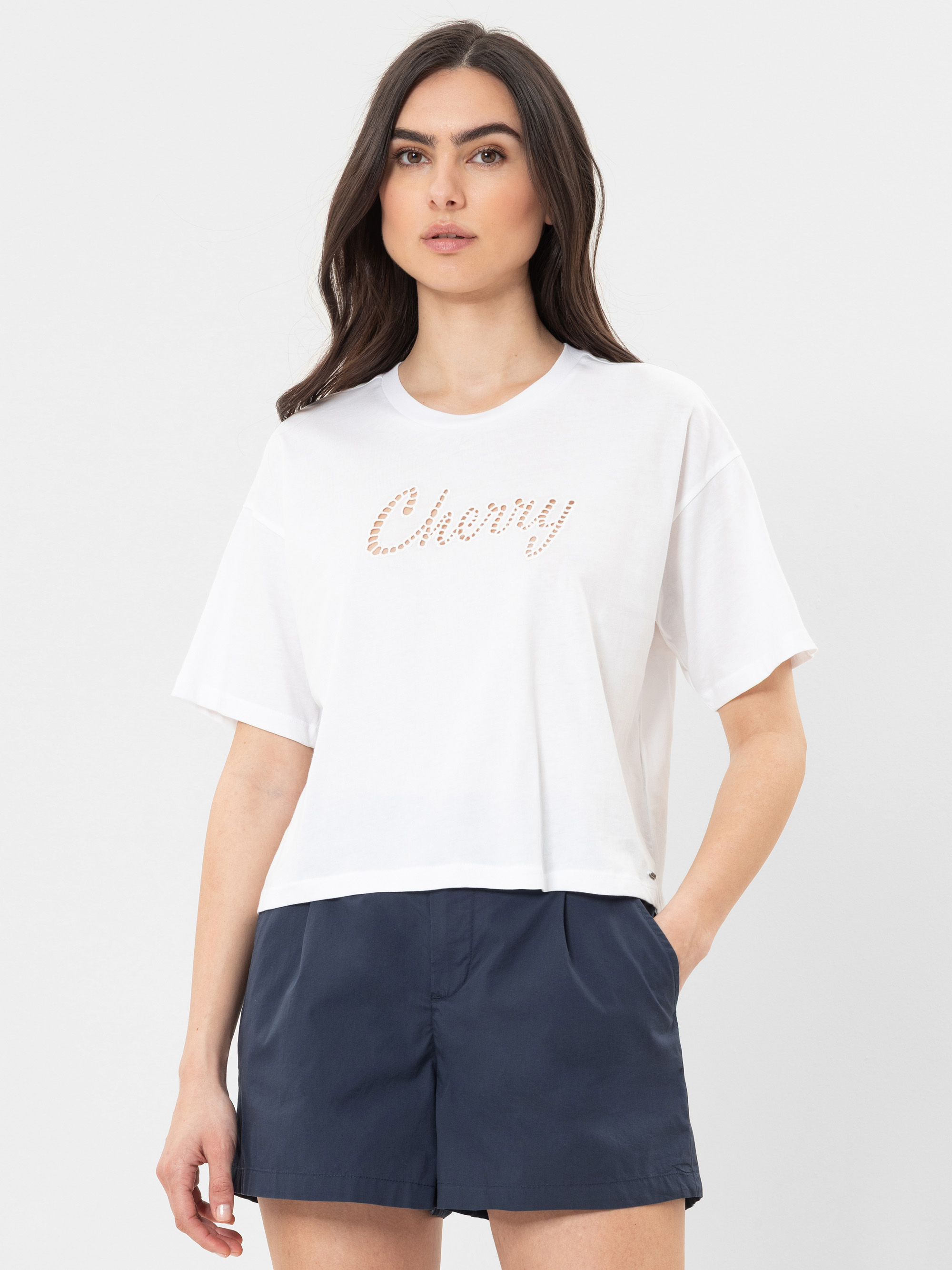 Eine Frau mit langen dunklen Haaren trägt ein weites weißes T-Shirt mit der Aufschrift Cherry und marineblaue Shorts vor einem schlichten hellen Hintergrund.