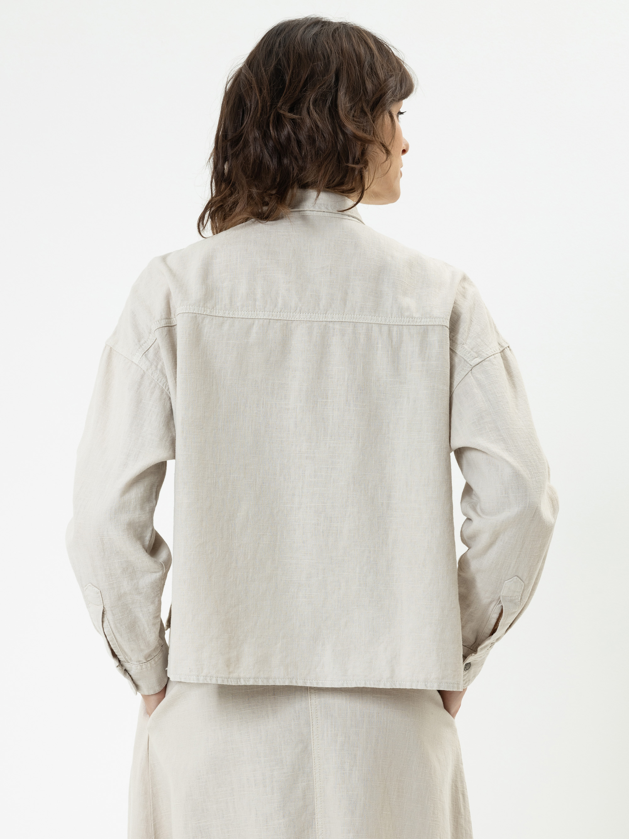 Eine Frau mit gewelltem braunem Haar ist von hinten zu sehen. Sie trägt ein hellbeiges, langärmeliges Hemd und einen passenden Rock. Sie steht vor einem schlichten weißen Hintergrund.