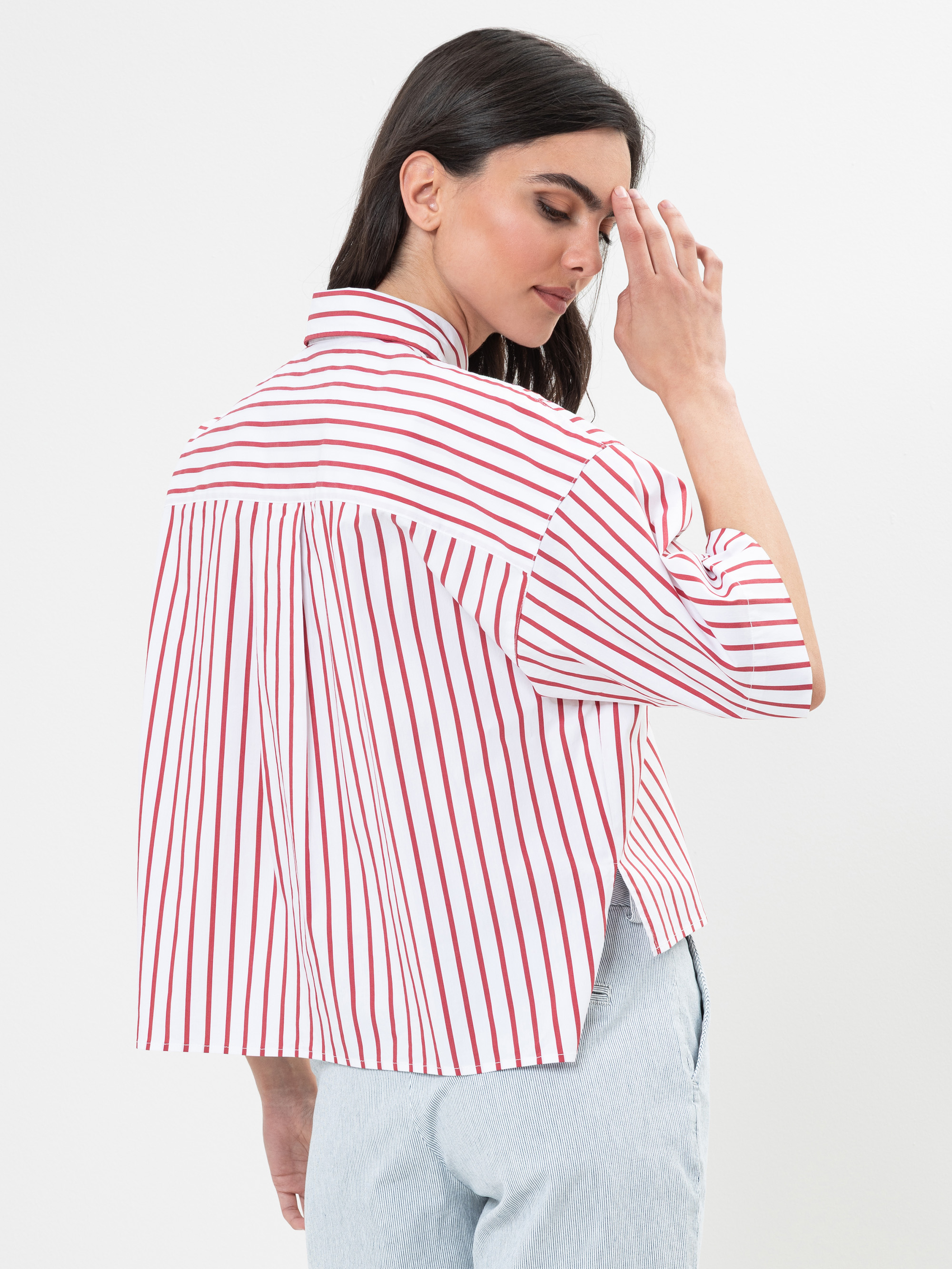 Eine Frau mit dunklem Haar, die ein weißes Hemd mit roten Längsstreifen und eine hellblaue Hose trägt, steht seitlich und berührt mit ihrer rechten Hand sanft ihr Gesicht vor einem schlichten weißen Hintergrund.