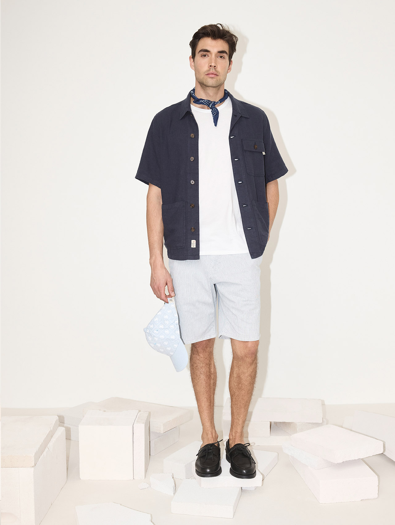 Ein Mann steht auf weißen Blöcken und trägt ein marineblaues kurzärmeliges Hemd über einem weißen T-Shirt, hell gestreifte Shorts, schwarze Schuhe und ein blaues Halstuch. In der linken Hand hält er eine hellblaue Mütze mit weißen Tupfen.