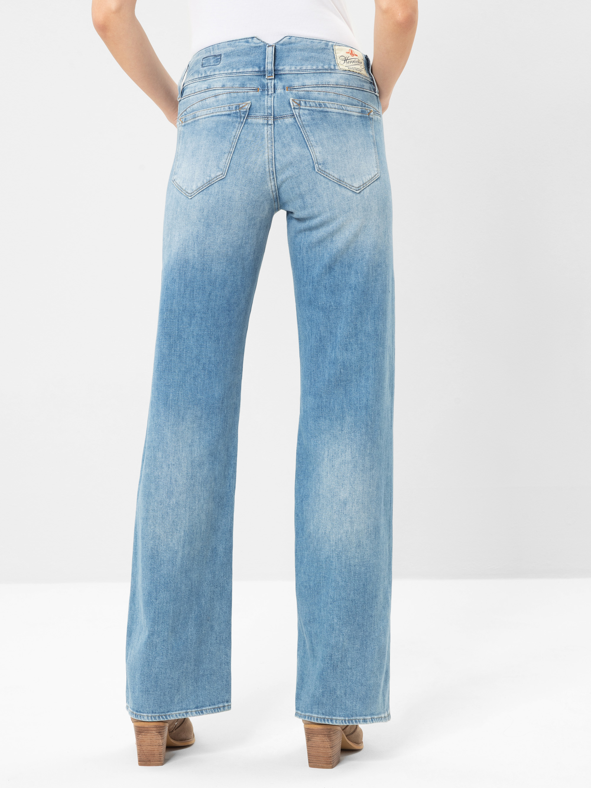 Eine Person steht mit dem Gesicht zur Wand, trägt eine hellblaue, weit geschnittene Jeans, ein weißes Oberteil und braune Schuhe mit Absätzen vor einem schlichten weißen Hintergrund.
