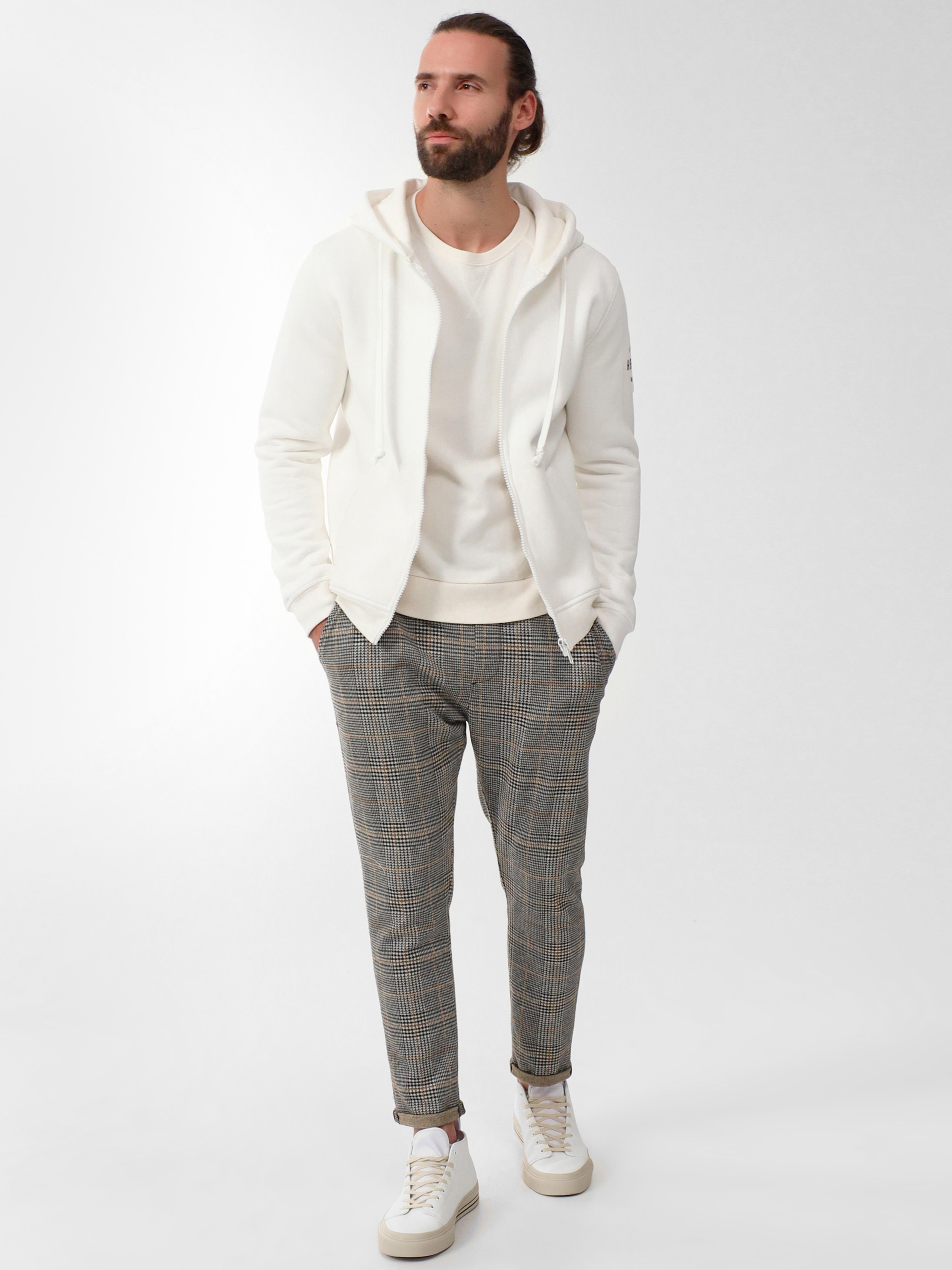Ein Mann mit Bart und langen, zurückgebundenen Haaren trägt einen weißen Kapuzenpullover, ein weißes T-Shirt, eine karierte Hose und weiße Turnschuhe. Er steht mit den Händen in den Taschen vor einem schlichten hellen Hintergrund.