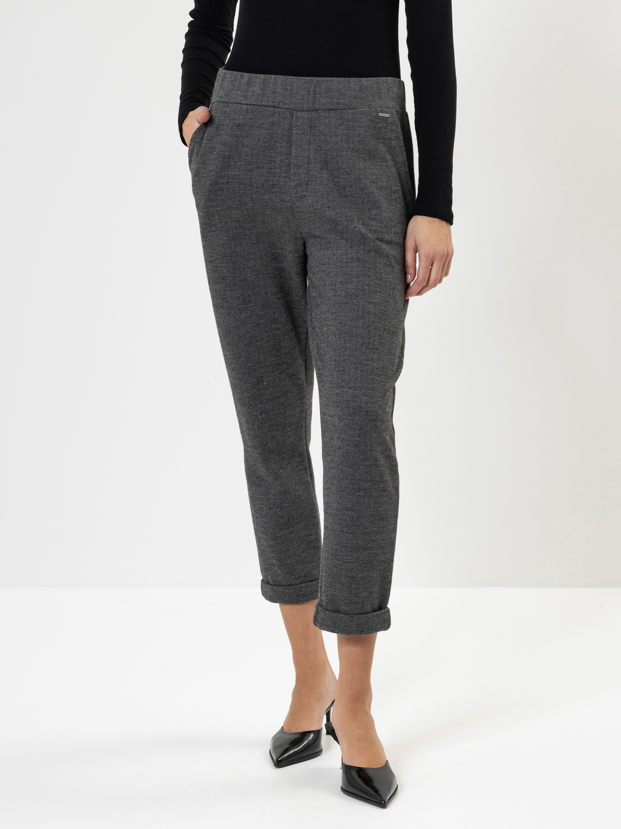 Eine Person, die eine graue, knöchellange Hose mit Manschetten und Händen in den Taschen, ein schwarzes, langärmeliges Oberteil und schwarze, spitz zulaufende Absätze trägt, steht vor einem schlichten weißen Hintergrund.