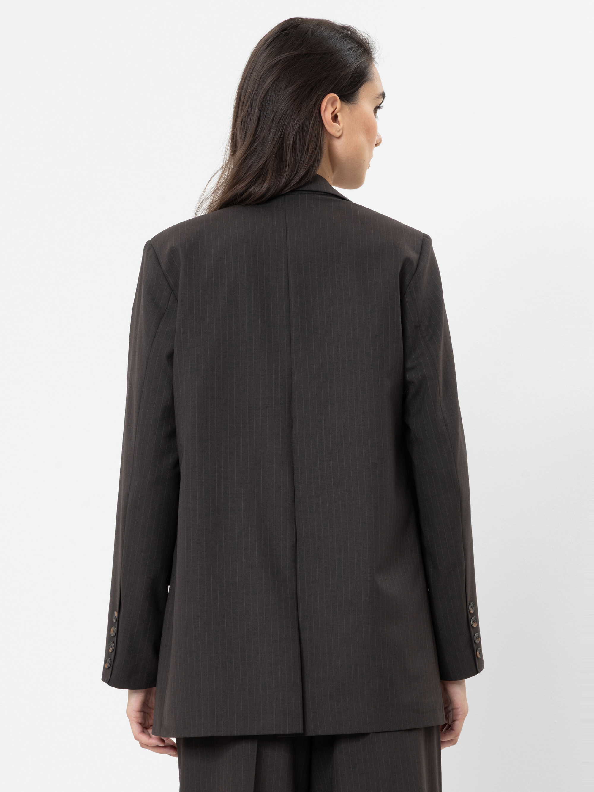 Eine Frau mit langen dunklen Haaren ist von hinten zu sehen; sie trägt eine locker sitzende dunkelbraune Anzugjacke und eine dazu passende Hose und steht vor einem schlichten weißen Hintergrund.