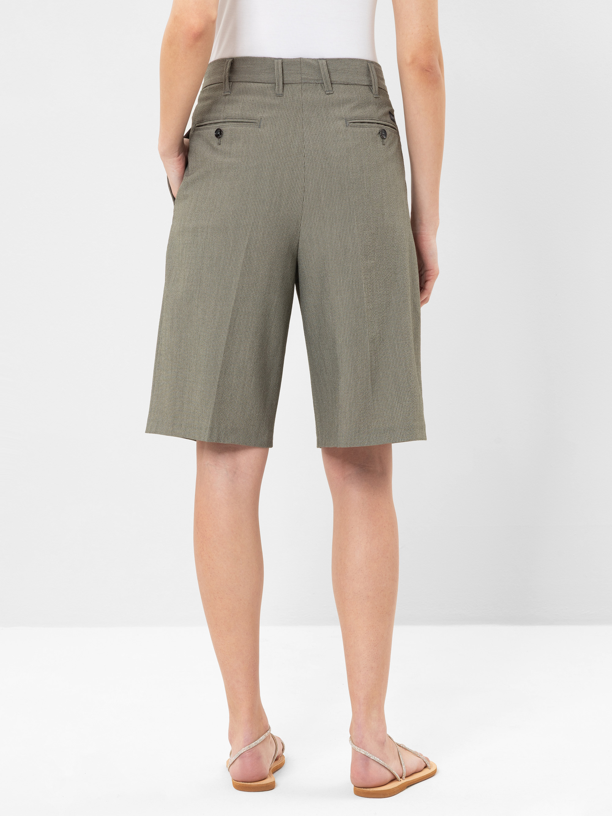 Eine Person, die ein weißes Oberteil, olivgrüne, knielange, maßgeschneiderte Shorts mit Gesäßtaschen und Knöpfen sowie hellbraune Sandalen trägt, steht vor einem schlichten weißen Hintergrund.