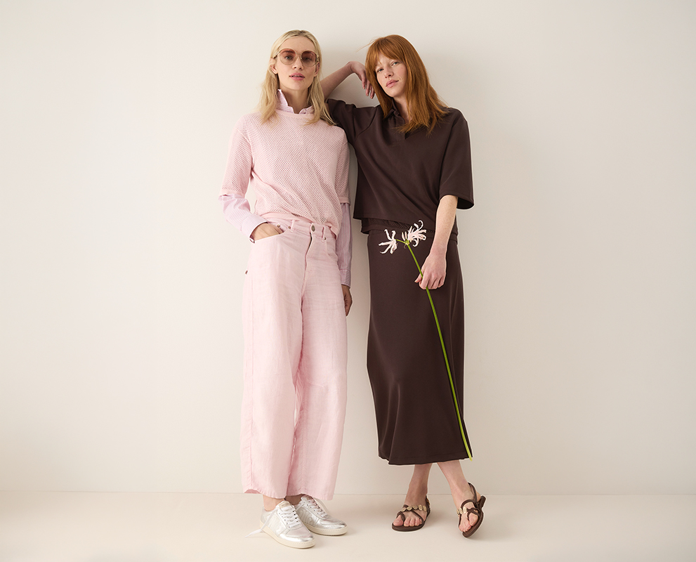 Zwei Frauen stehen zusammen vor einem schlichten Hintergrund; die eine trägt eine hellrosa Hose, ein passendes Hemd und eine Sonnenbrille, die andere ein braunes Kleid und hält eine weiße Blume. Beide sehen entspannt und stilvoll aus.