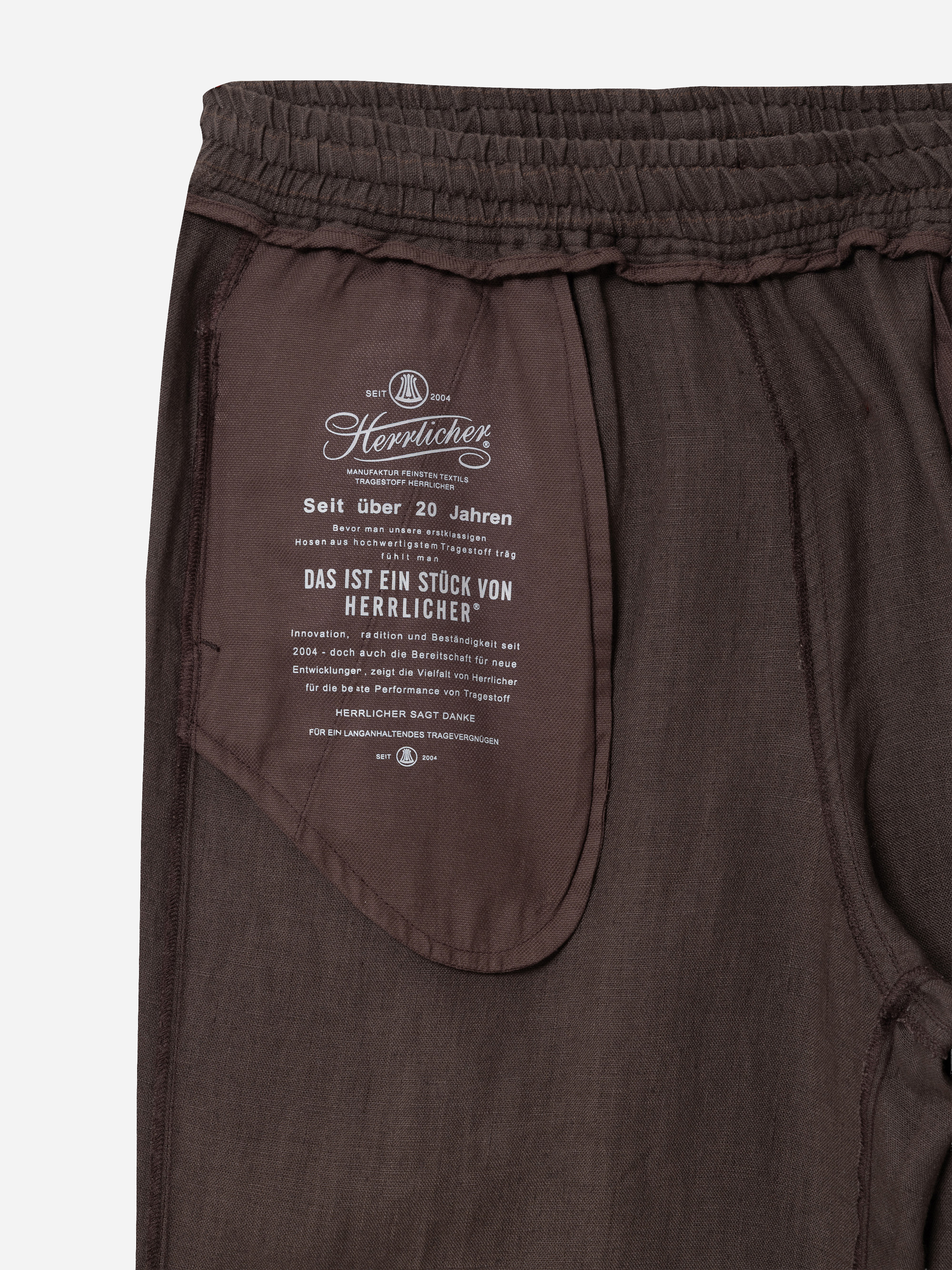 Nahaufnahme der Innenseite einer braunen Hose, die eine Tasche mit weißem Aufdruck und Branding von Herrlicher zeigt. Der Text ist in deutscher Sprache und enthält Informationen zur Qualität und zum Unternehmen. Der Bund ist elastisch.