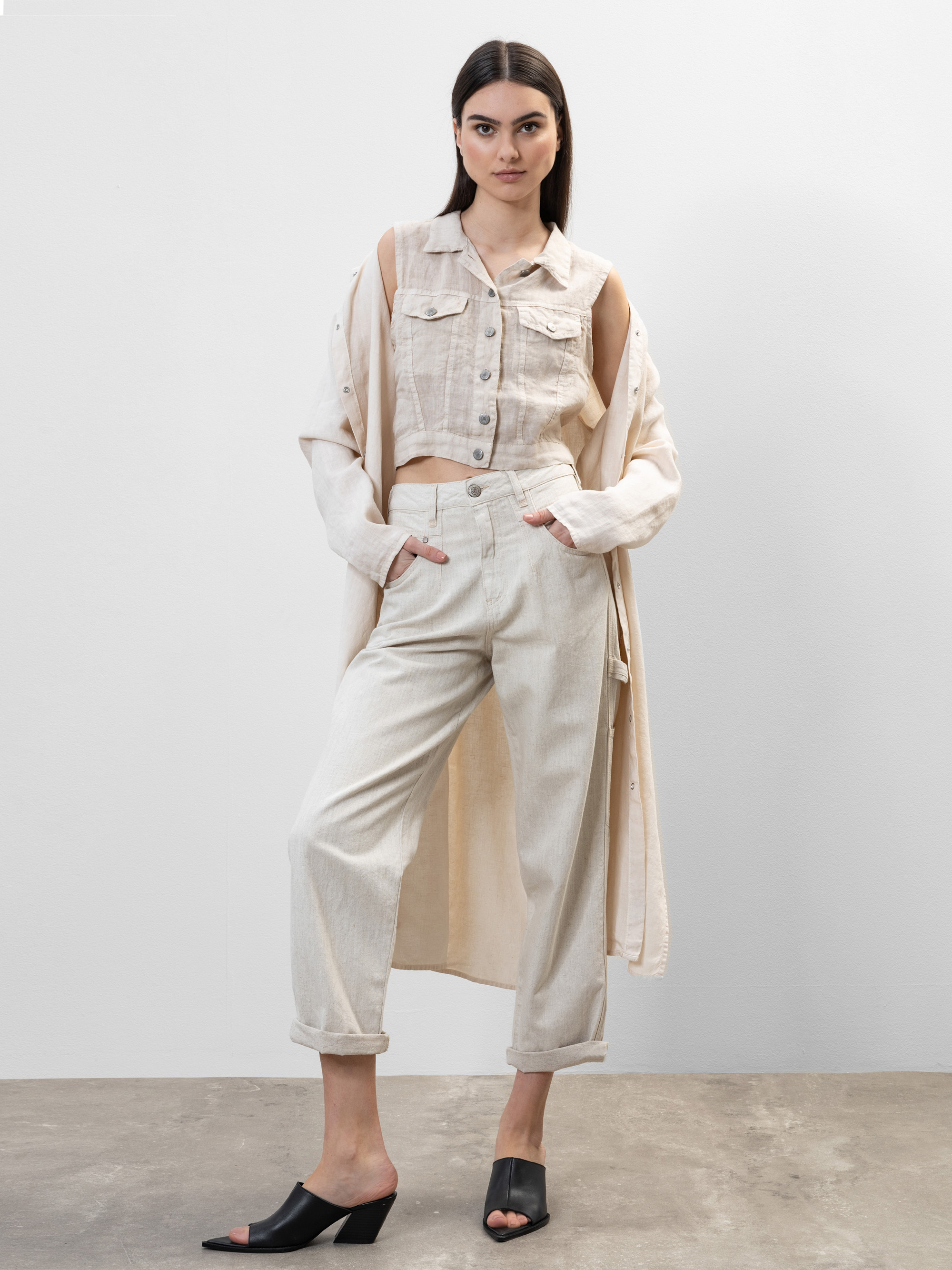 Eine Frau steht vor einem unifarbenen Hintergrund und trägt ein helles, ärmelloses, geknöpftes Oberteil, eine dazu passende hochtaillierte Hose mit gerollten Bündchen, eine lange, fließende Jacke und schwarze, hochhackige Sandalen. Sie hat langes, gl
