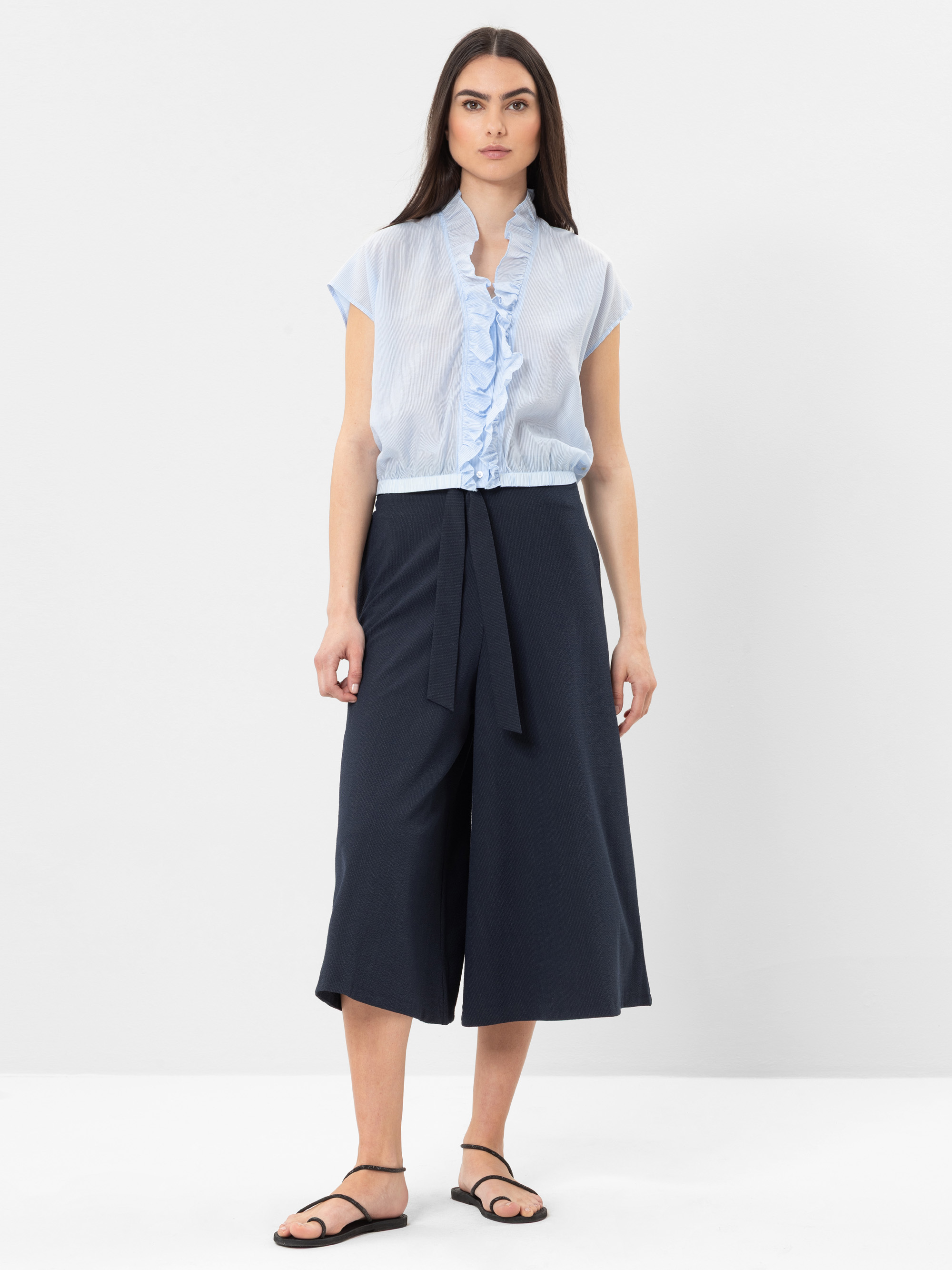 Eine Frau steht vor einem schlichten weißen Hintergrund und trägt eine hellblaue Rüschenbluse, eine marineblaue Culottes mit weitem Bein, die in der Taille gebunden ist, und schwarze Sandalen. Sie hat langes glattes Haar und einen neutralen Gesichtsaus