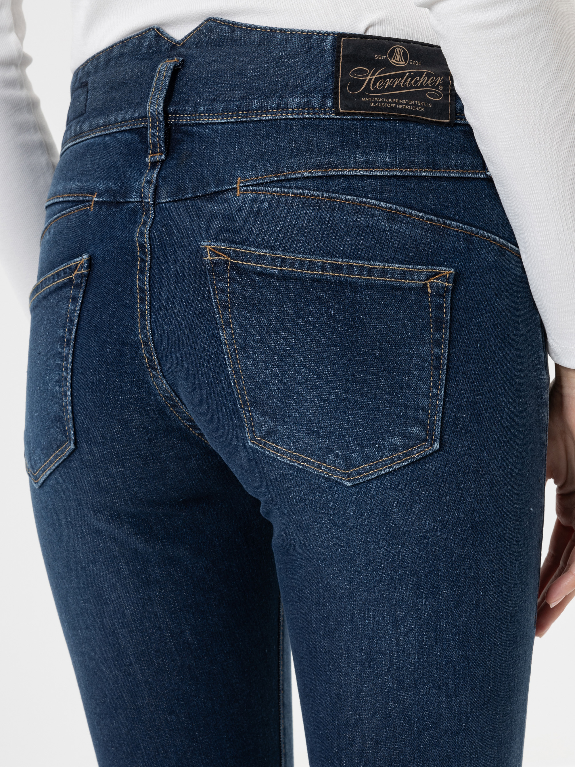 Nahaufnahme einer Person, die eine dunkelblaue Jeans und ein weißes Oberteil trägt, wobei die Gesäßtaschen und ein schwarzer Markenaufnäher aus Leder am Bund zu sehen sind. Das Bild konzentriert sich auf die Passform und die Details der Nähte der Je