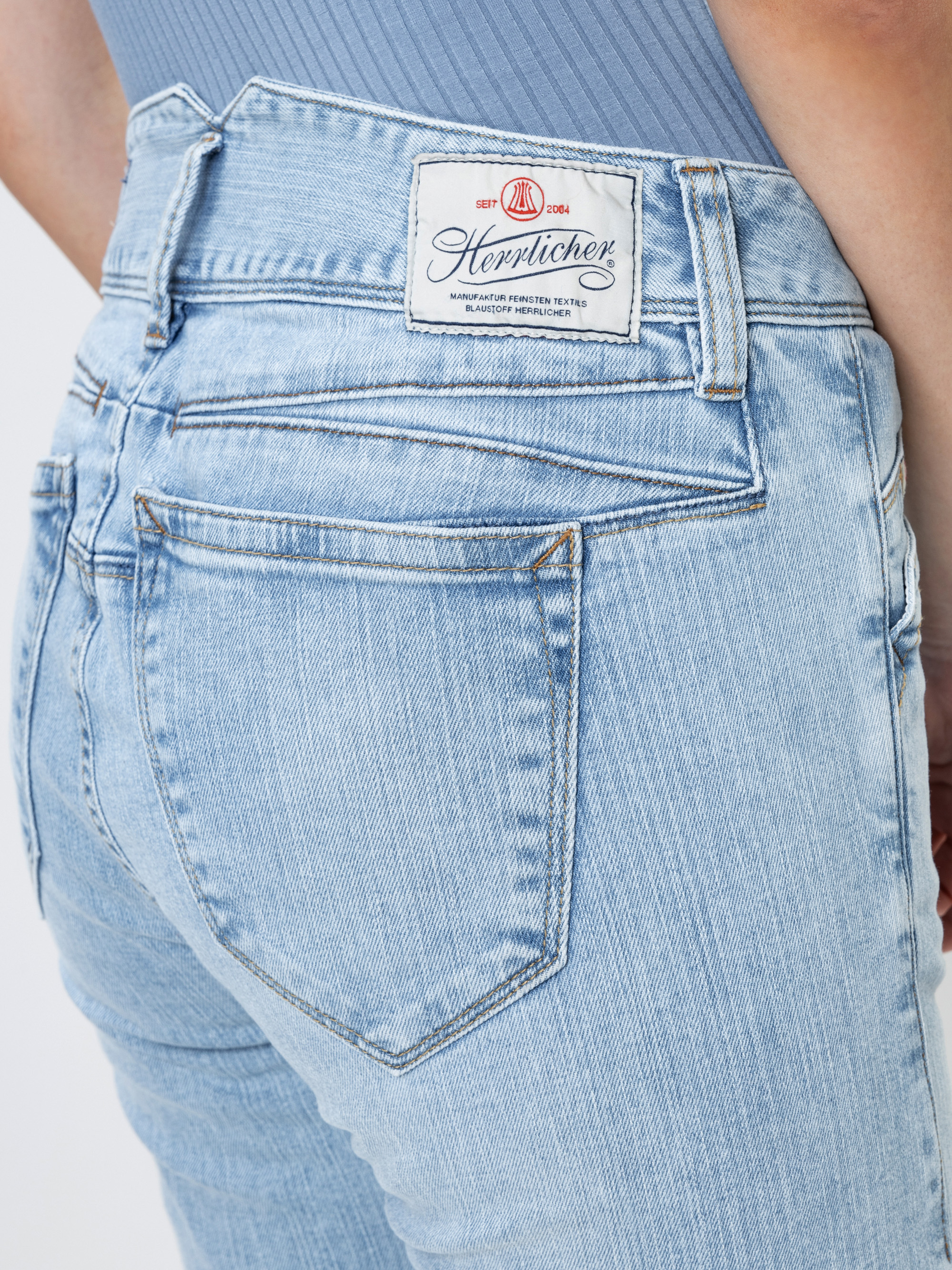 Nahaufnahme einer Person, die eine hellblaue Jeans mit einem Herrlicher-Markenaufnäher am Bund trägt, wobei die Gesäßtasche und ein Teil eines blauen Rippenhemds zu sehen sind.