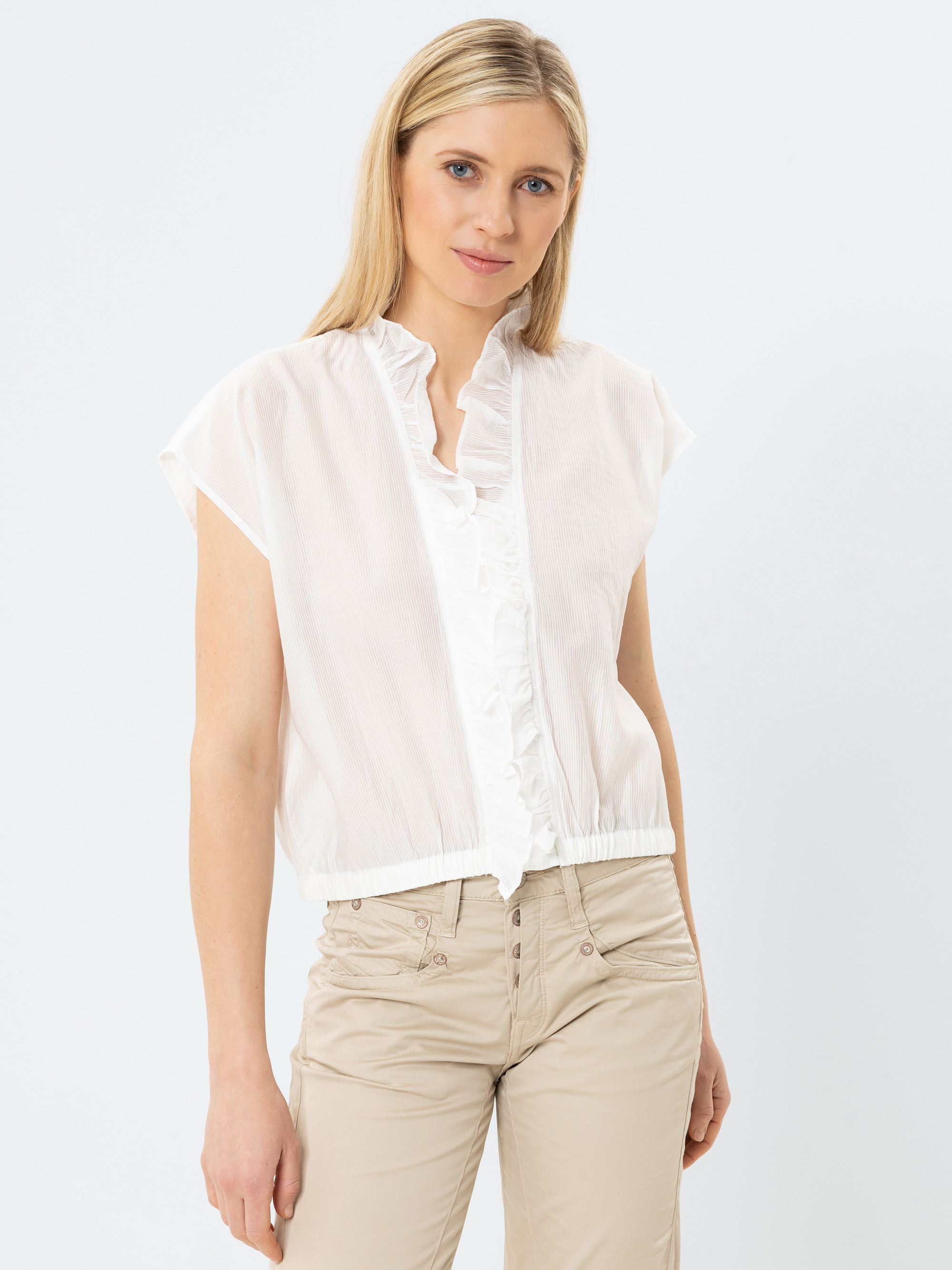 Eine Frau mit langem blondem Haar trägt eine weiße kurzärmelige Bluse mit Rüschenkragen und Knöpfen, gepaart mit einer hellbeigen Hose, und steht vor einem schlichten weißen Hintergrund.