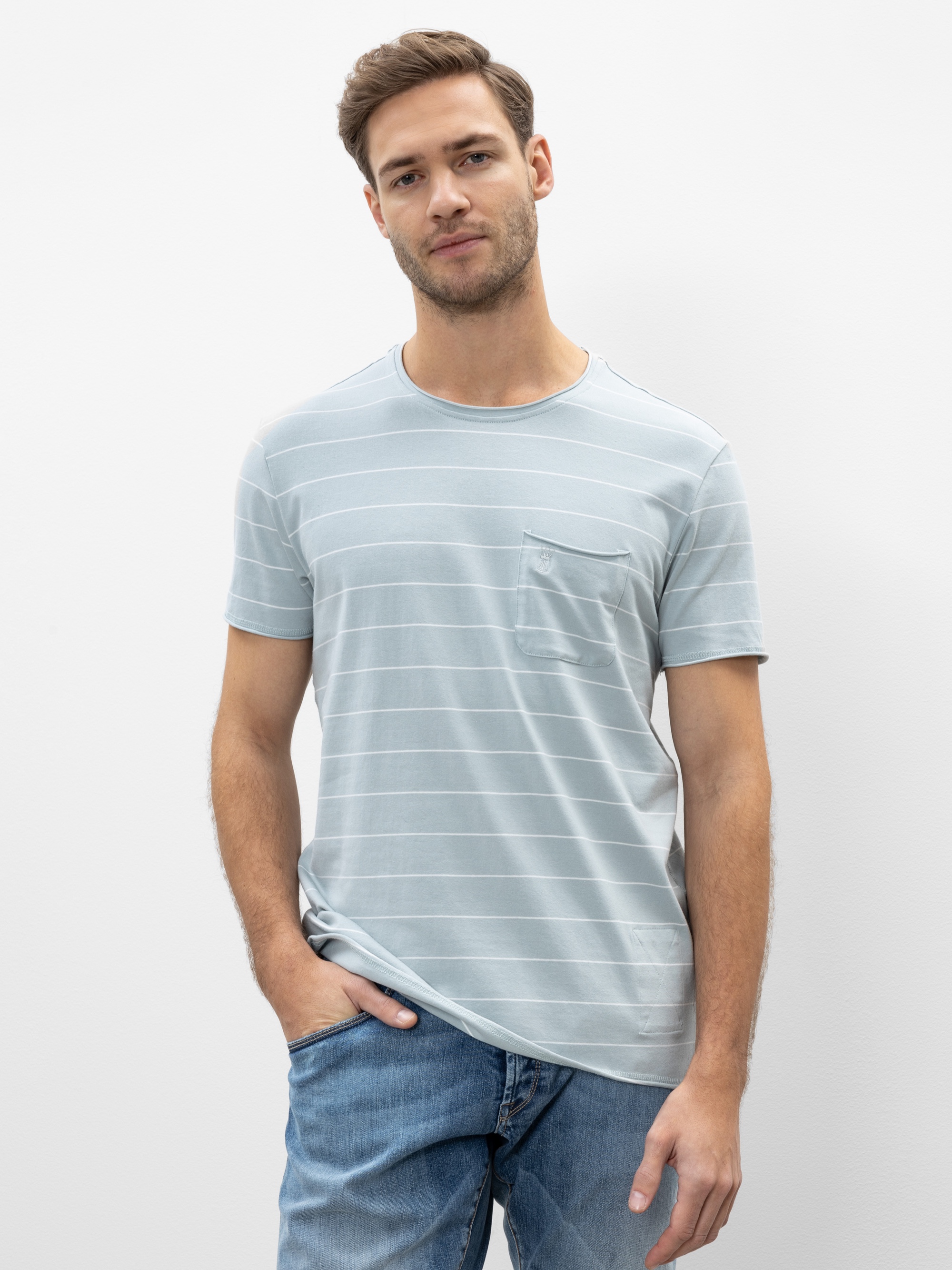 Ein Mann mit kurzen braunen Haaren und gestutztem Bart trägt ein hellblaues, kurzärmeliges, gestreiftes T-Shirt mit einer Brusttasche und blaue Jeans. Er steht vor einem schlichten weißen Hintergrund und hat eine Hand in der Tasche.