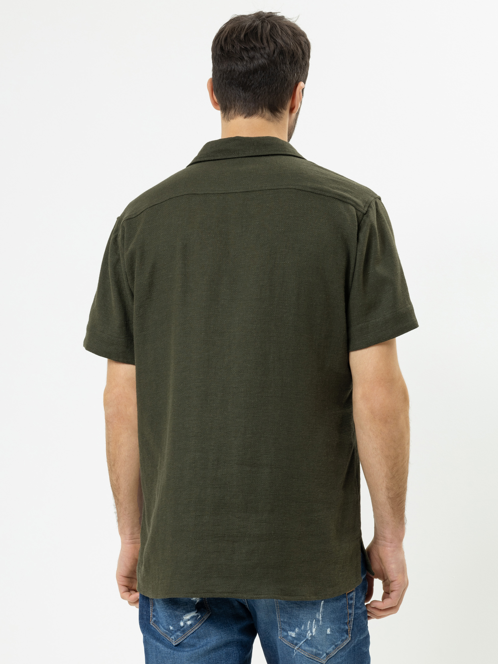 Ein Mann steht mit dem Rücken zur Kamera und trägt ein kurzärmeliges, dunkelgrünes Hemd und blaue Jeans mit leichten Gebrauchsspuren. Der Hintergrund ist einfarbig weiß.