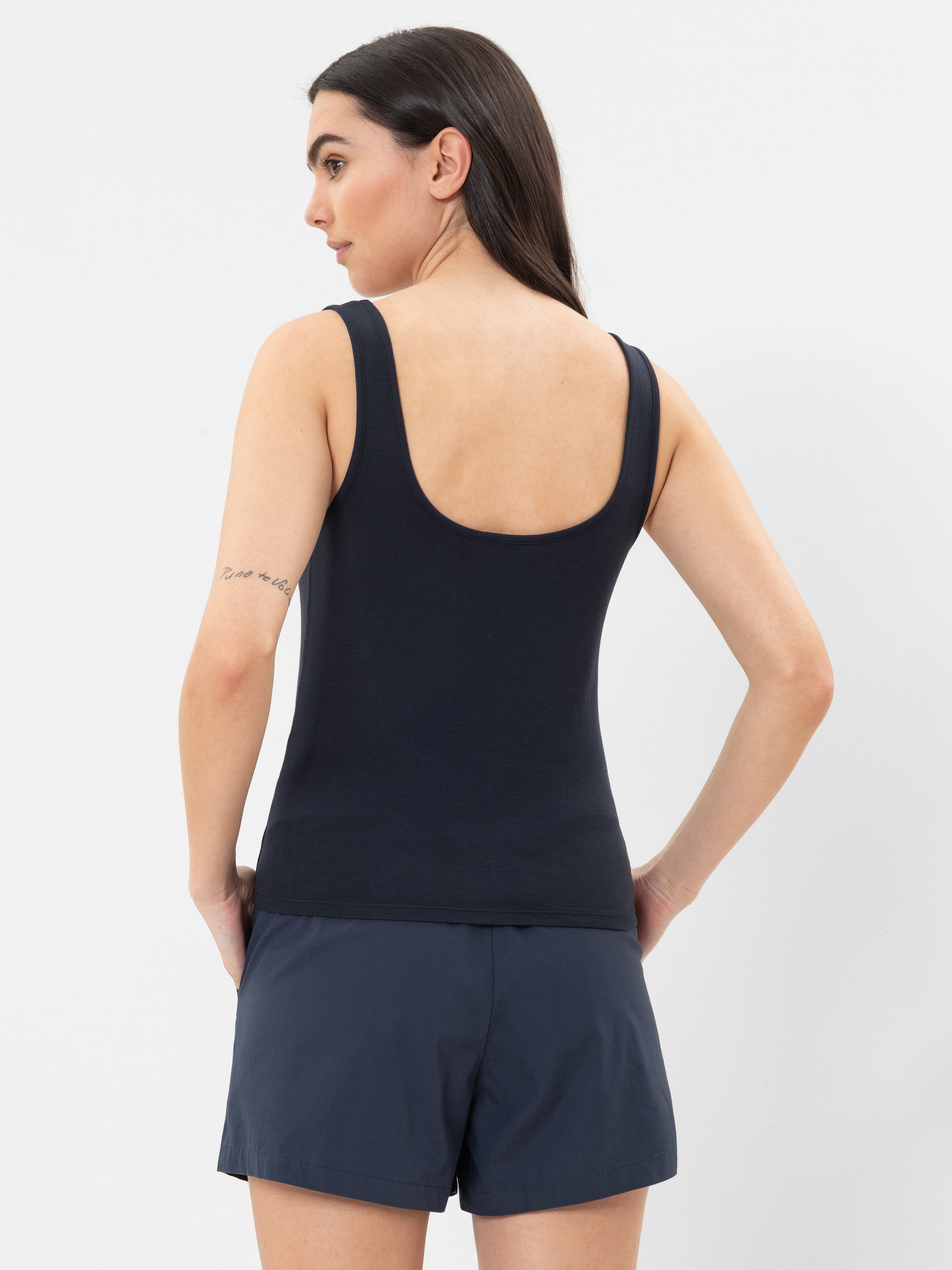 Eine Frau mit langen dunklen Haaren steht mit dem Rücken zur Kamera, trägt ein ärmelloses schwarzes Oberteil und dunkle Shorts. Sie hat eine sichtbare Tätowierung auf ihrem linken Oberarm und steht vor einem schlichten weißen Hintergrund.