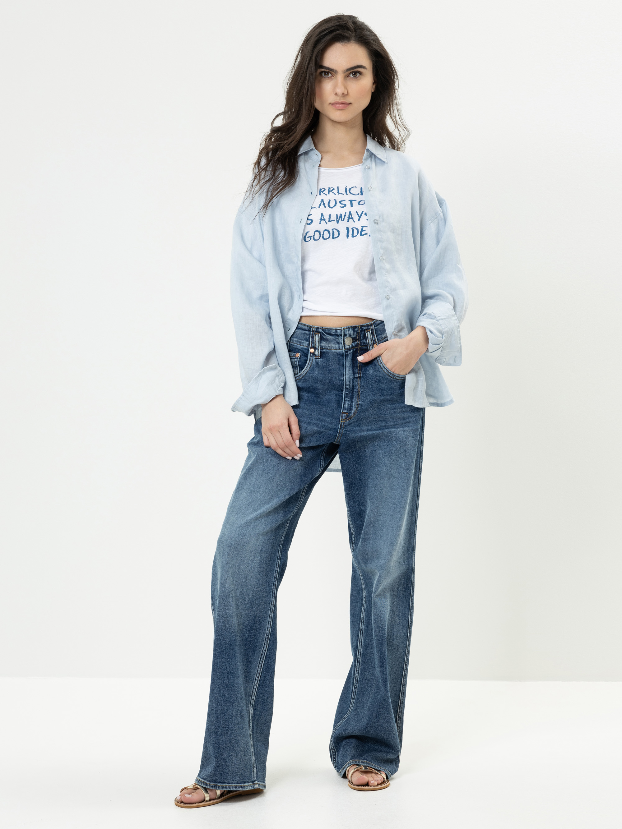 Eine junge Frau mit langen dunklen Haaren steht vor einem schlichten weißen Hintergrund. Sie trägt ein hellblaues Button-up-Hemd über einem weißen grafischen T-Shirt, weite Blue Jeans und braune Sandalen. Sie posiert mit einer Hand in ihrer Tasche.